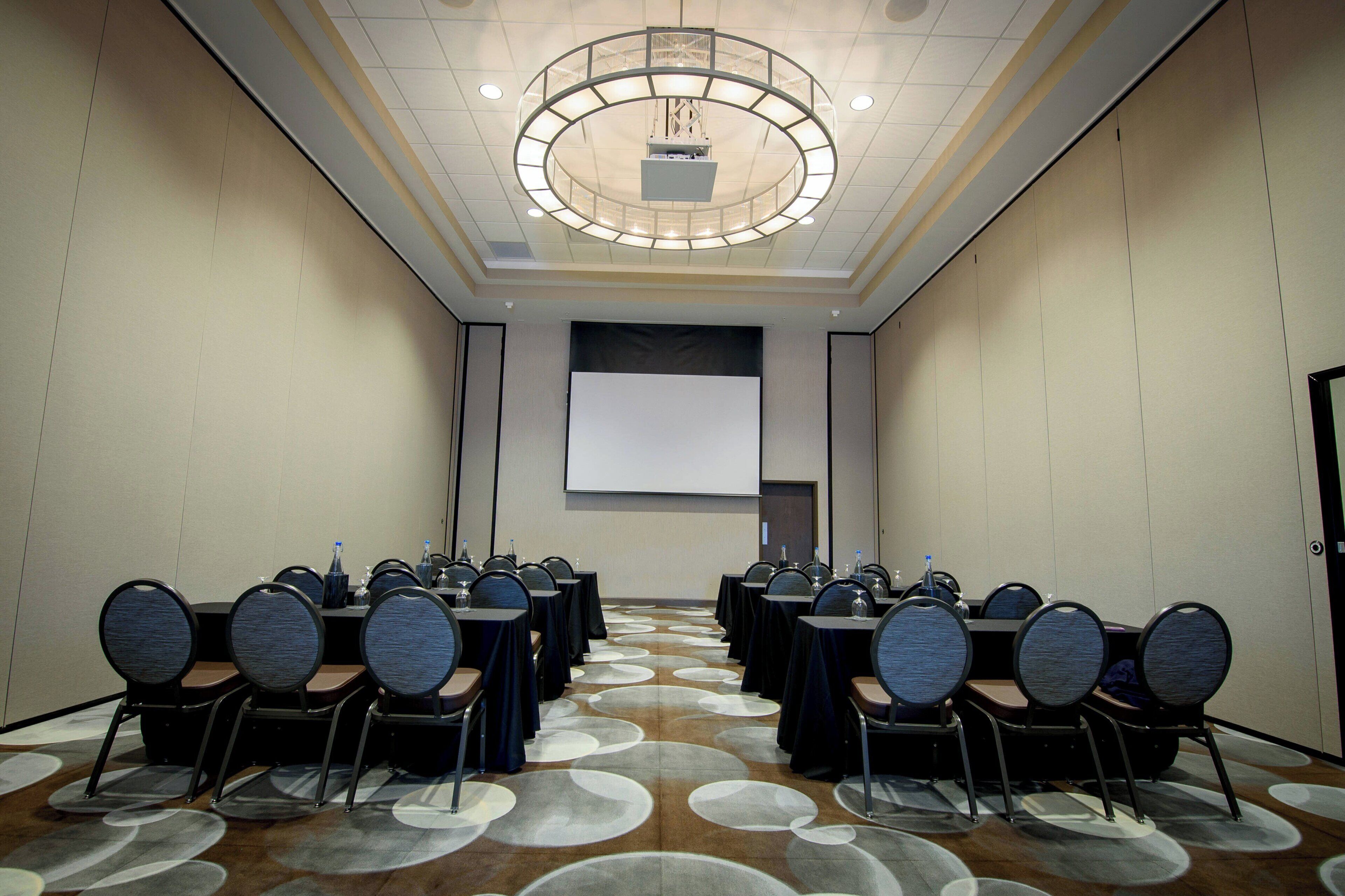 A conference room with rows of tables and chairs, a projector screen, and a large screen on the wall.