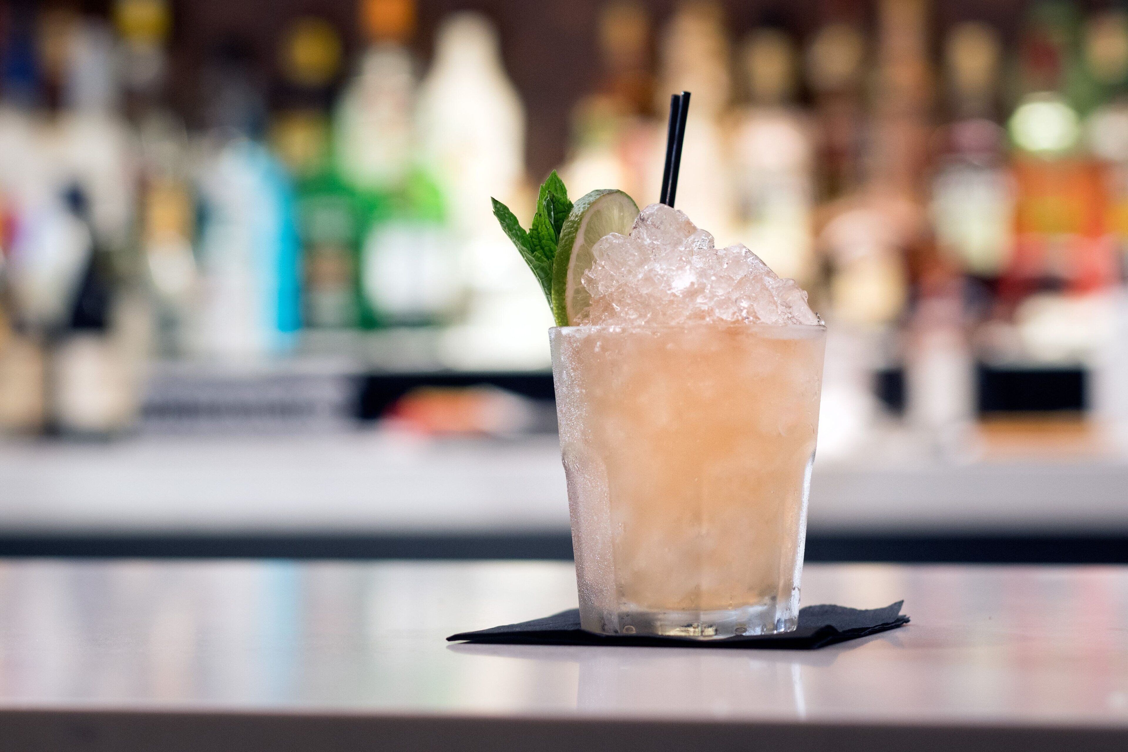 Eine Bar-Szene mit einem Glas gekühltem Getränk, garniert mit Minze und einer Limettenscheibe, Eiswürfeln und einem Strohhalm.