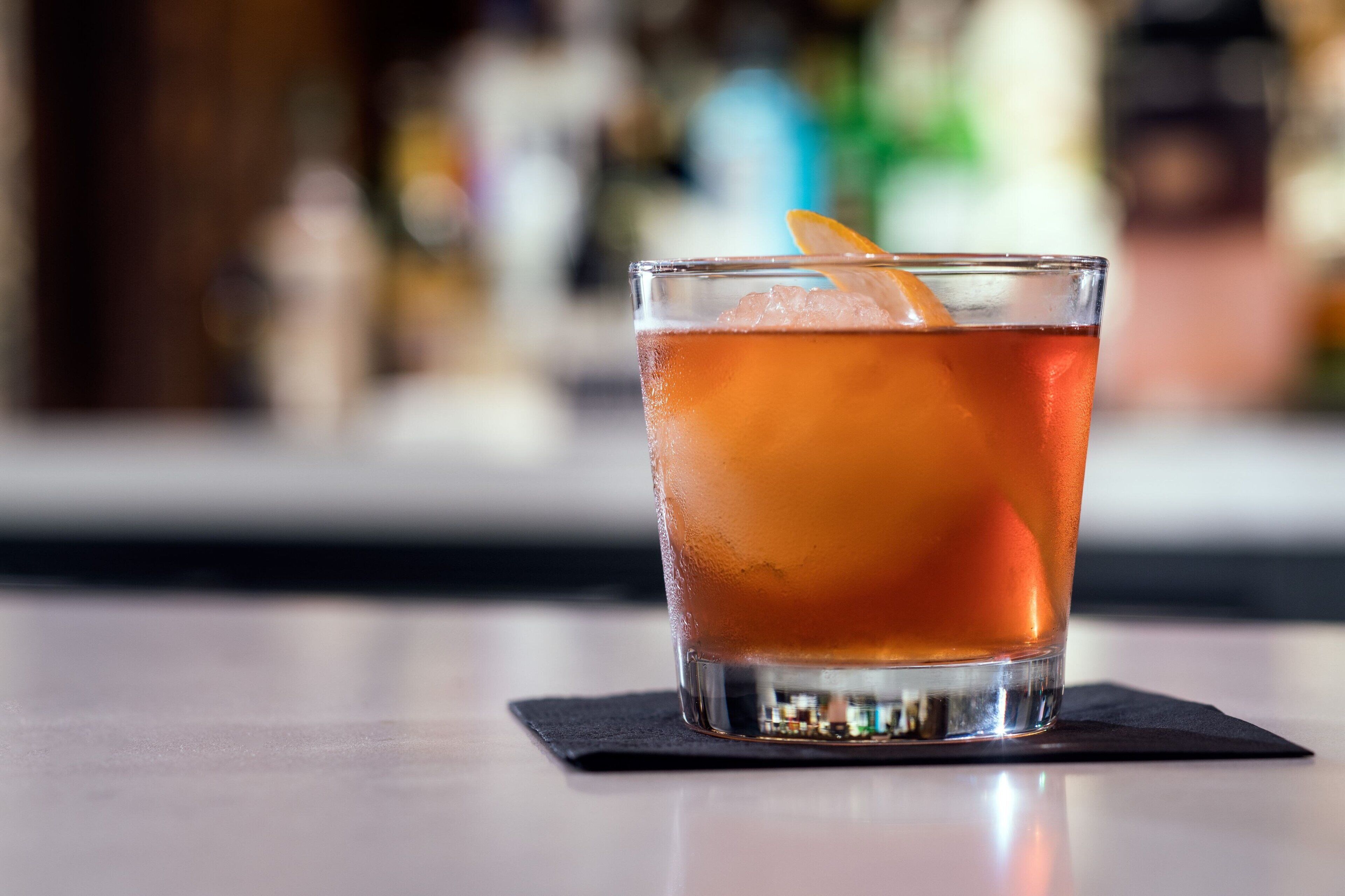 Ein Glas bernsteinfarbenes Getränk mit Orangenscheibe auf einem schwarzen Untersetzer.