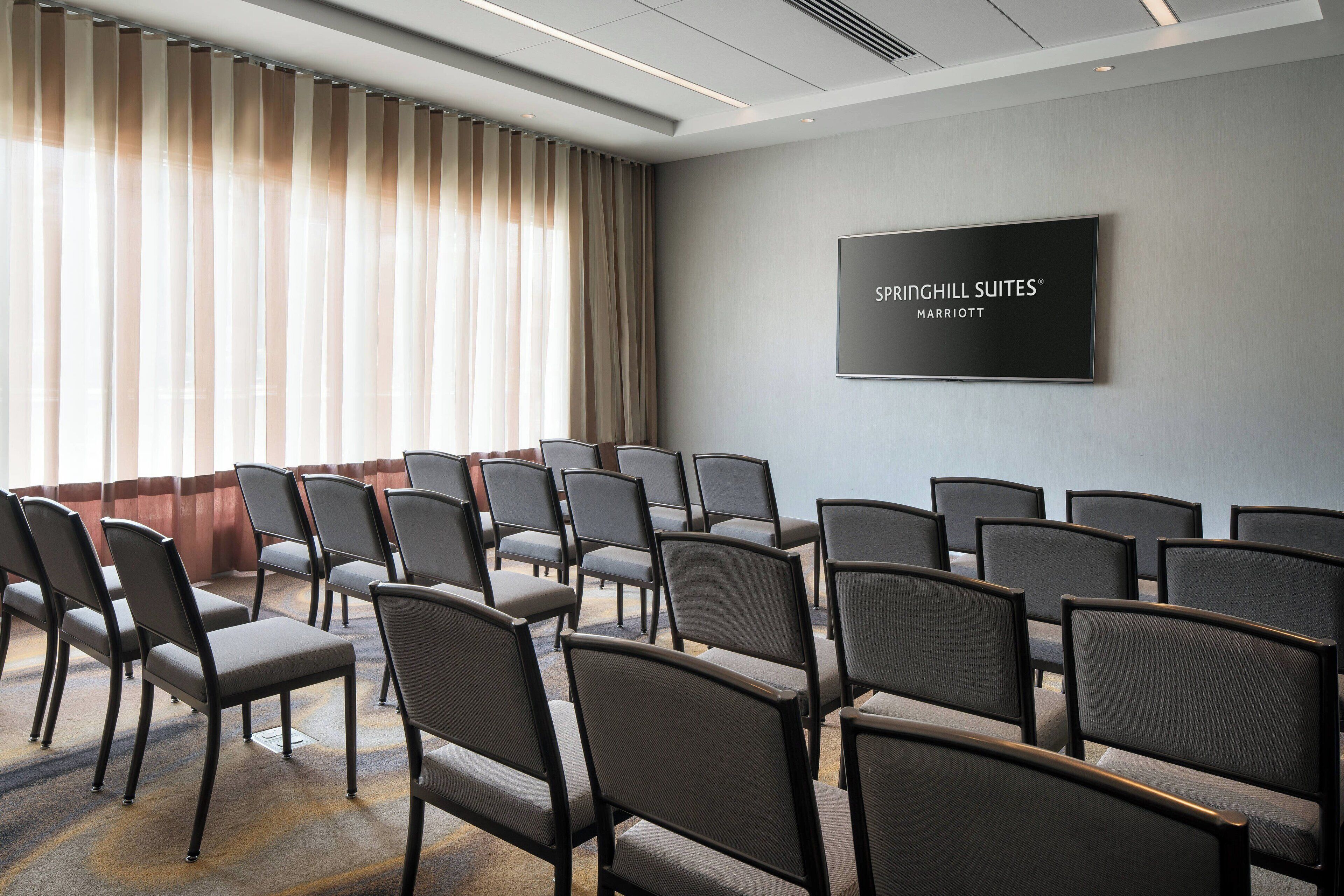 A conference room with rows of chairs, a large screen displaying 'Springhill Suites® Marriott', and a large window with curtains.