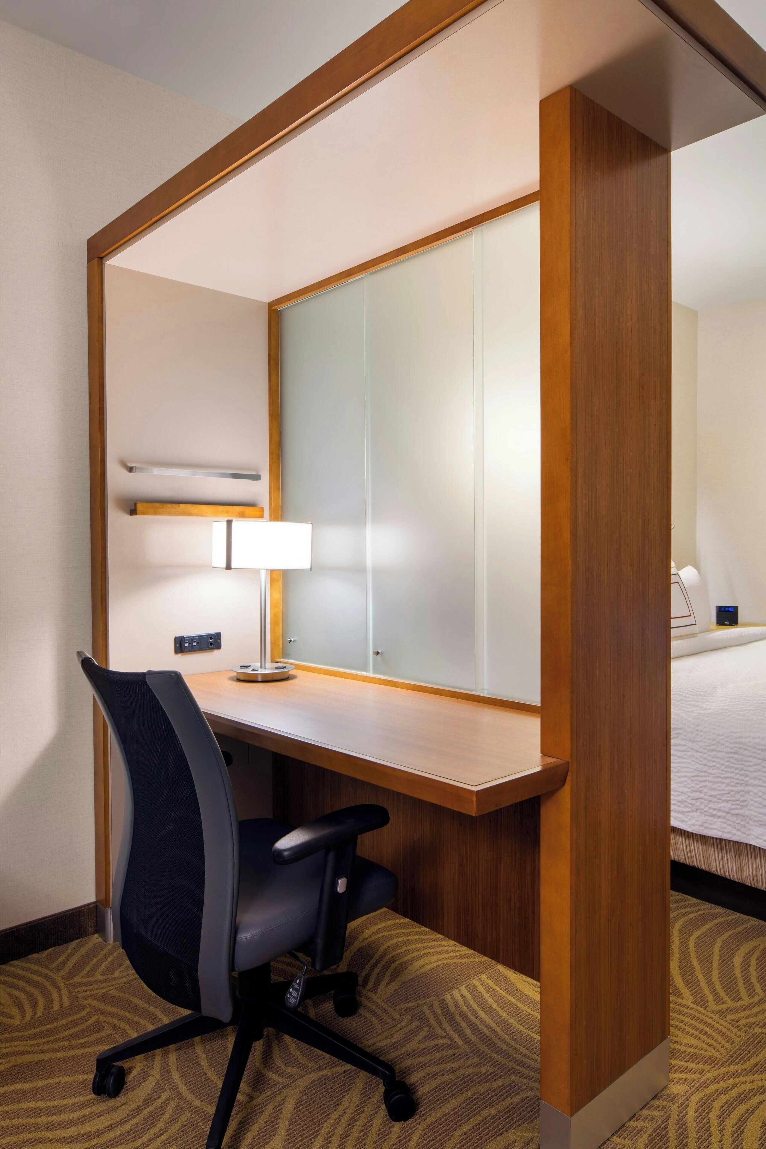 Habitación de hotel con un escritorio de madera, una silla de oficina negra, una lámpara, una cama y un espejo grande.