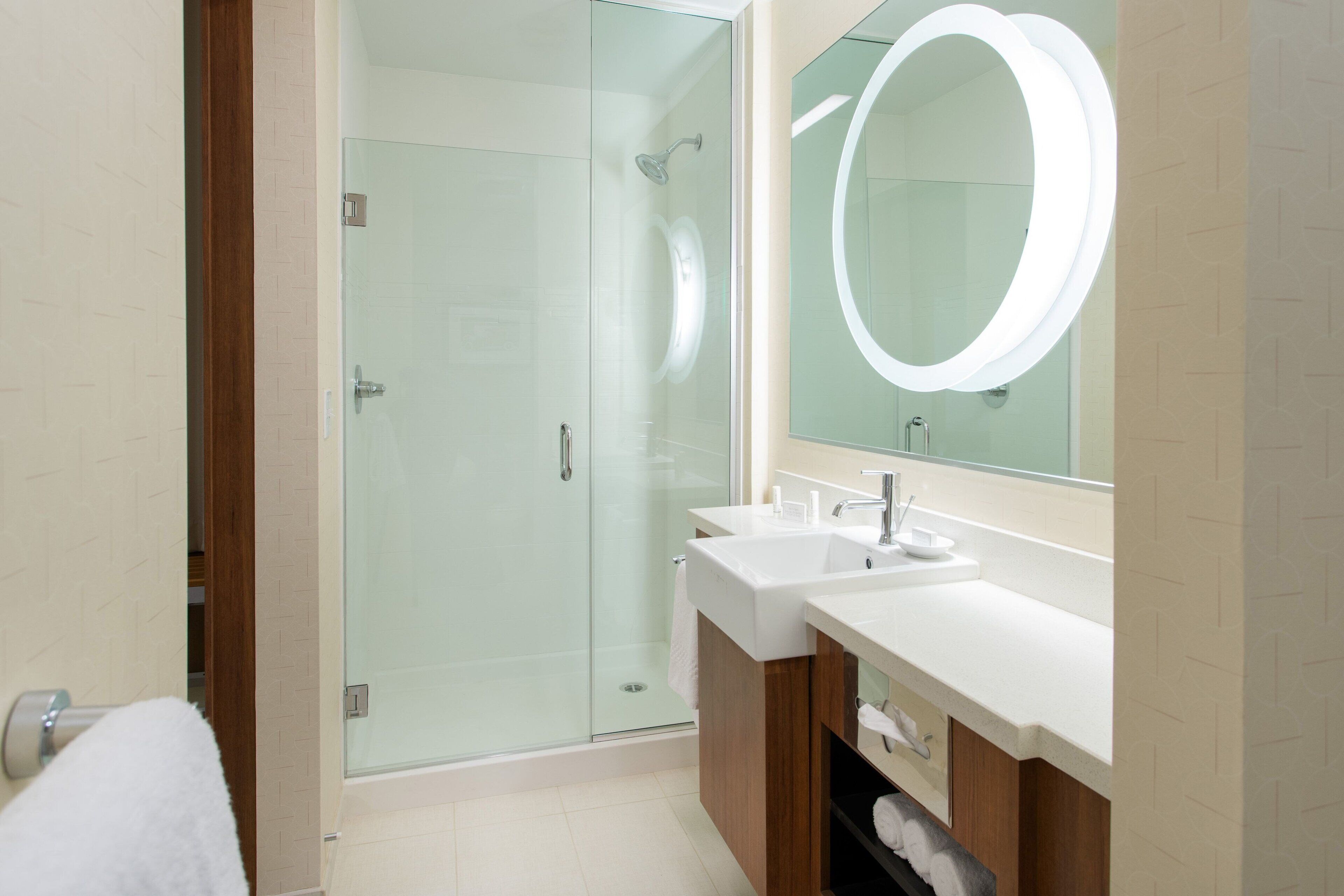 Un baño moderno con ducha envuelta en vidrio, dos lavamanos y un espejo grande con luz circular.