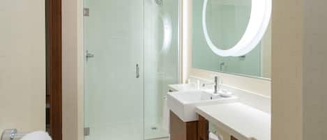 Un baño moderno con ducha envuelta en vidrio, dos lavamanos y un espejo grande con luz circular.