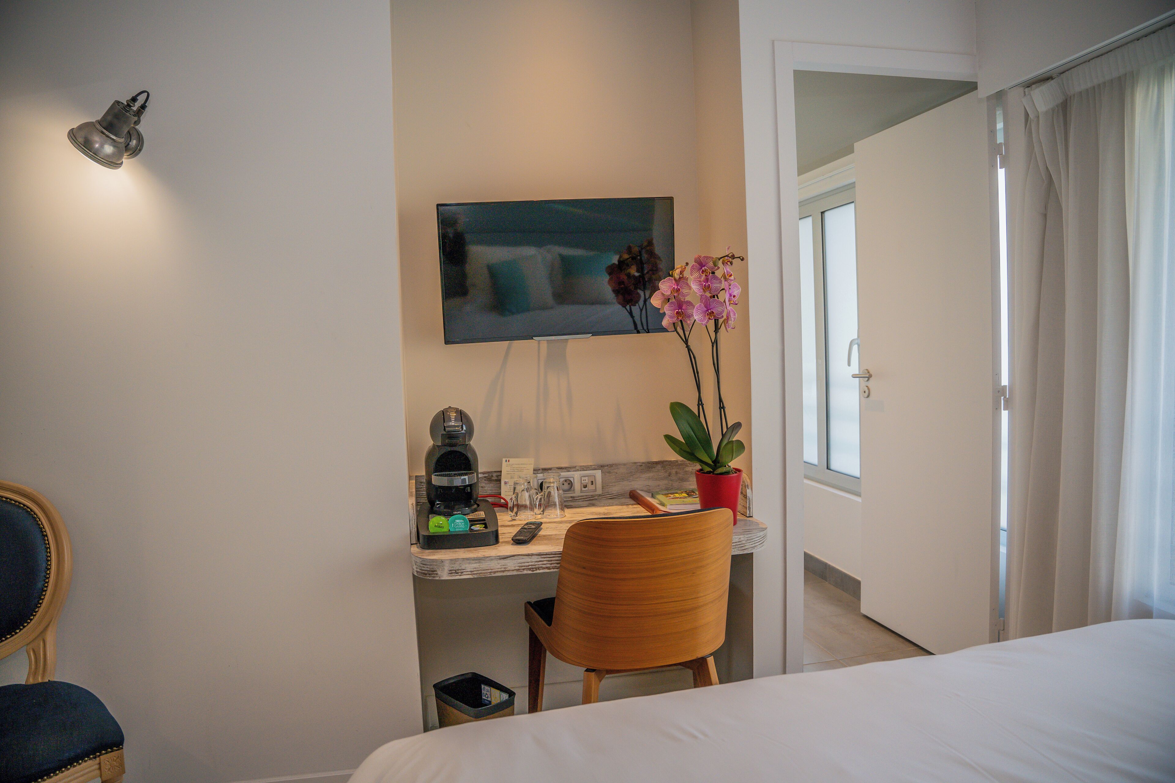 A hotel room with a bed, a desk with a coffee maker, a chair, a TV mounted on the wall, and a potted plant.