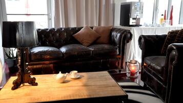 A dark leather sofa with two cushions, a wooden coffee table with a teapot and cups, a lamp, and a table with a coffee machine and cups.