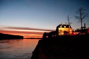 A building with a prominent roof and a lit sign, situated by a body of water at dusk. - Les 3 Lieux - Hôtel, Restaurant, Espace sensoriel (Les Ponts-de-Ce)