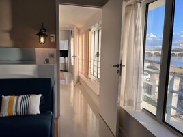 A modern hotel room with a large window, a sofa, and a hallway leading to a bathroom.