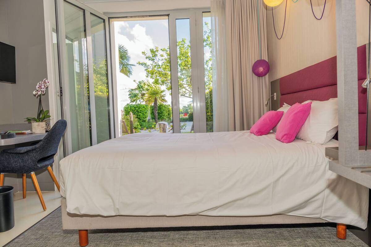 Habitación de hotel con una cama grande, un escritorio y una silla. Se ve follaje desde la ventana.