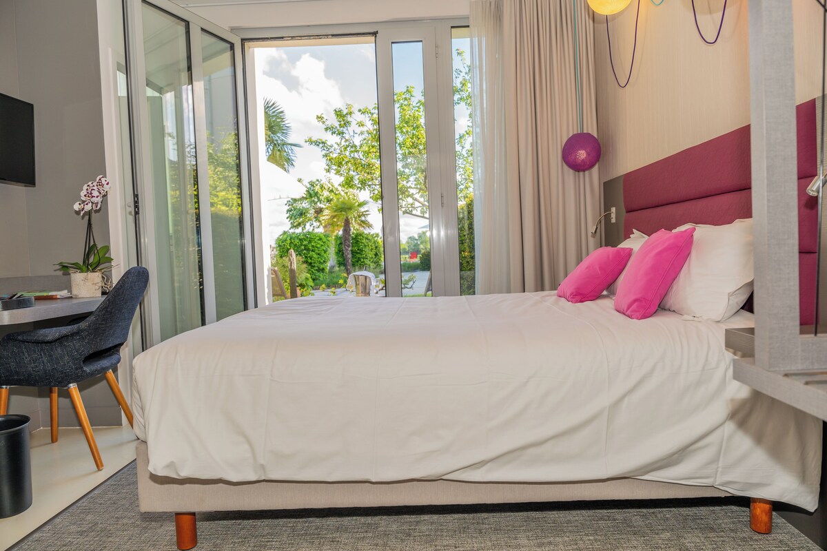 A hotel room with a large bed, a desk, and a chair. There is a view of greenery outside the window.