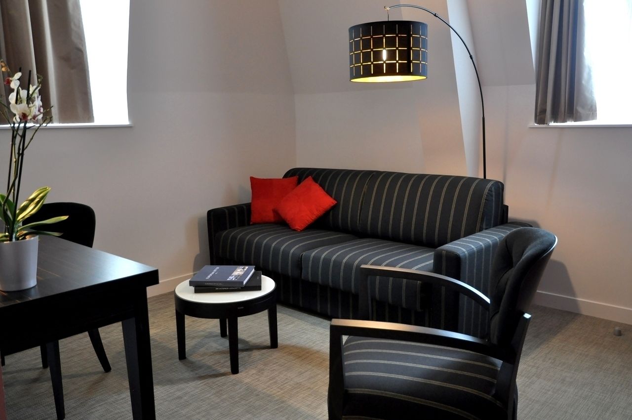 Een moderne woonkamer met een gestreept bankje, een rode kussen, een klein rond tafeltje, een vloerlamp en een potplant.