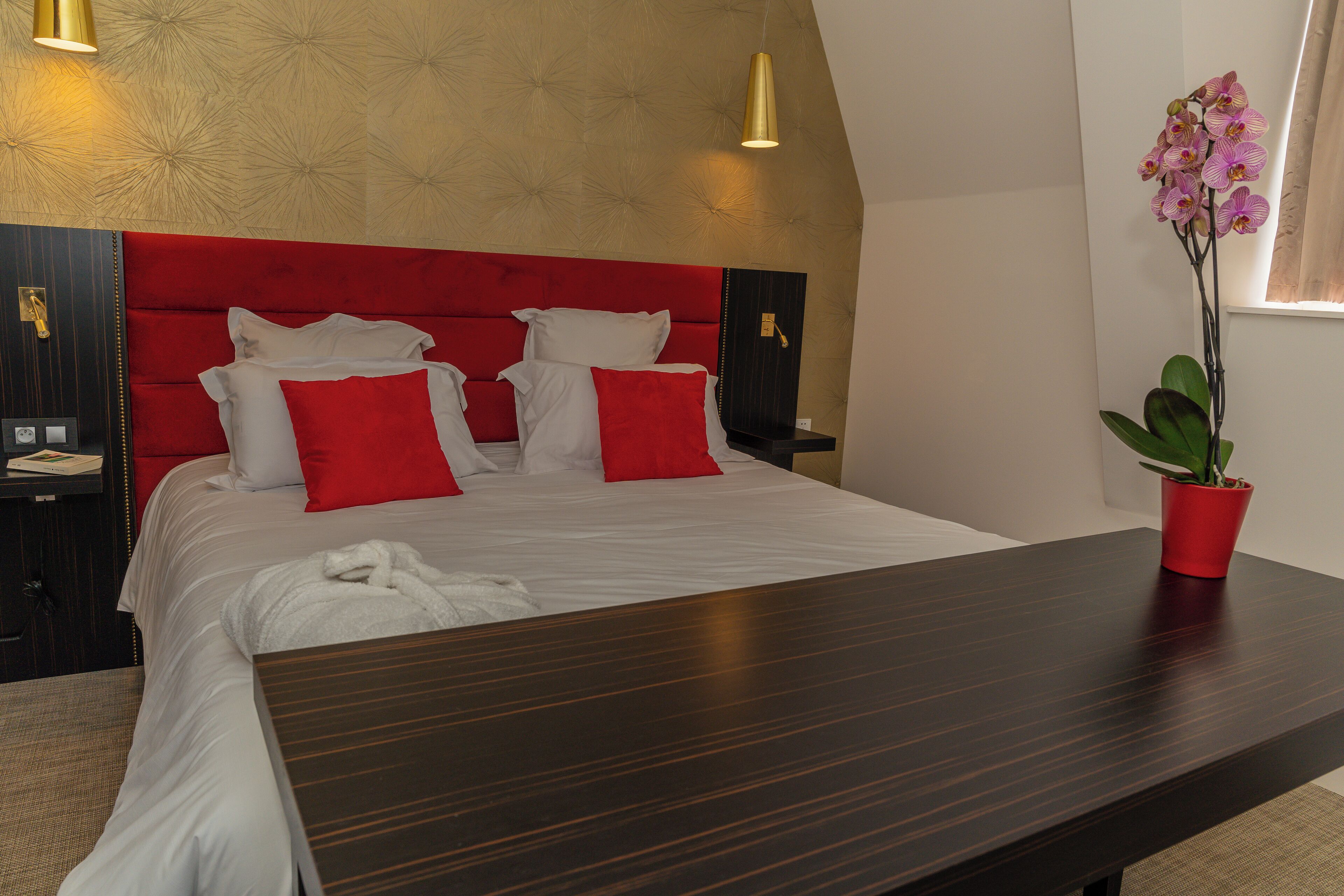 Habitación de hotel con una cama grande, ropa de cama roja y blanca, una mesita de noche de madera, una planta en maceta roja y una lámpara fijada a la pared.