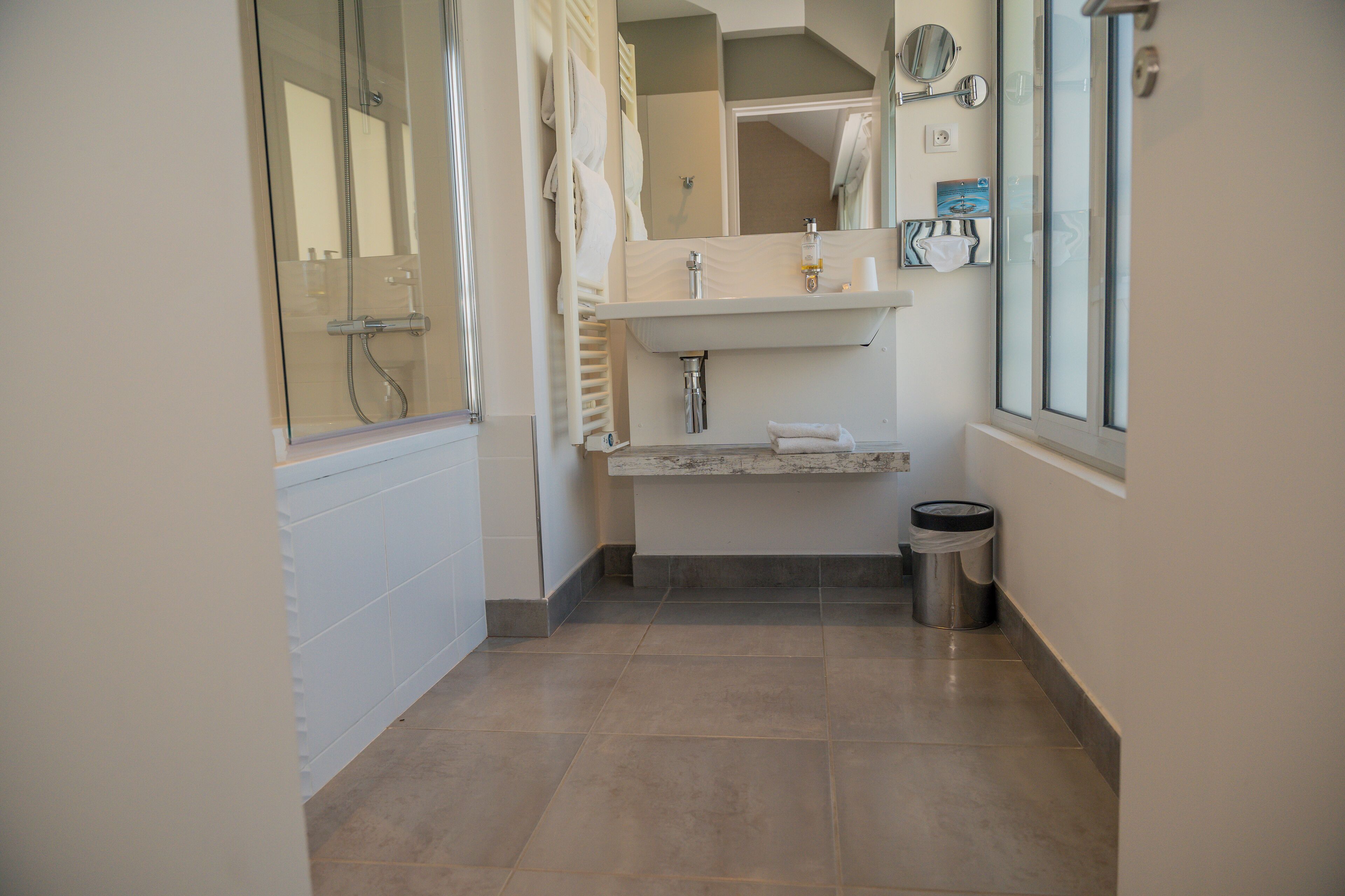 A bathroom with a glass-enclosed shower, a vanity with a mirror, and a trash can.