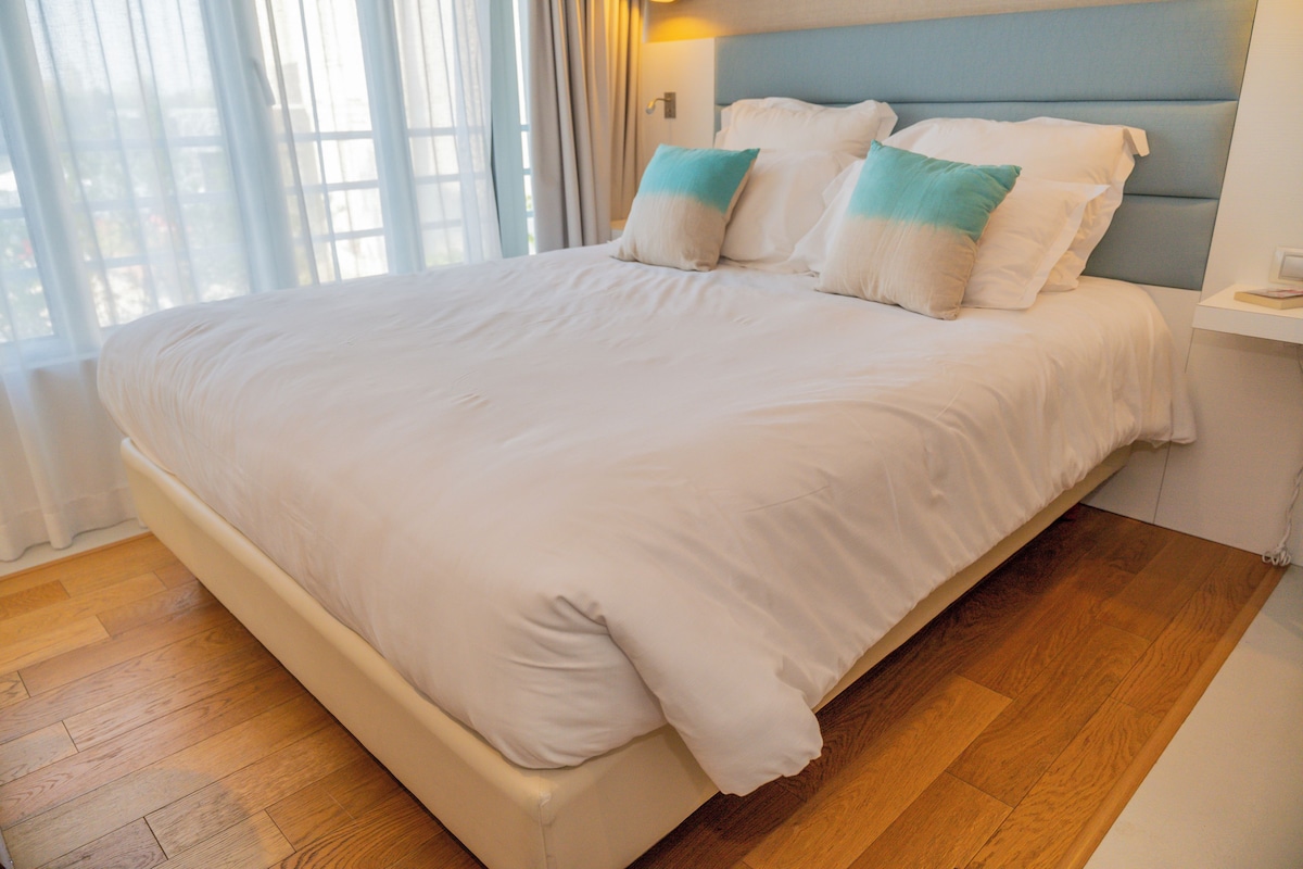 A large bed with white bedding and teal pillows, a wooden floor, and a nightstand with a lamp.
