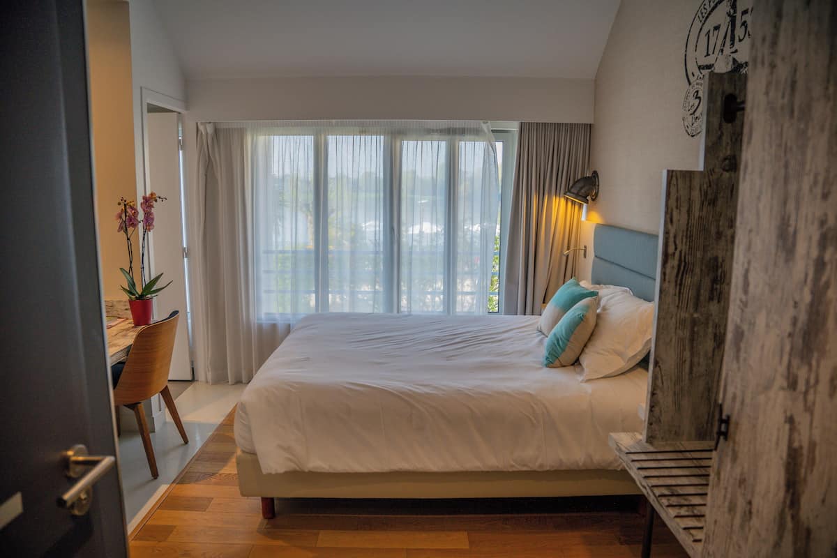 Un dormitorio con una cama grande, un escritorio con una silla, una planta en maceta y vistas al exterior a través de cortinas translúcidas.