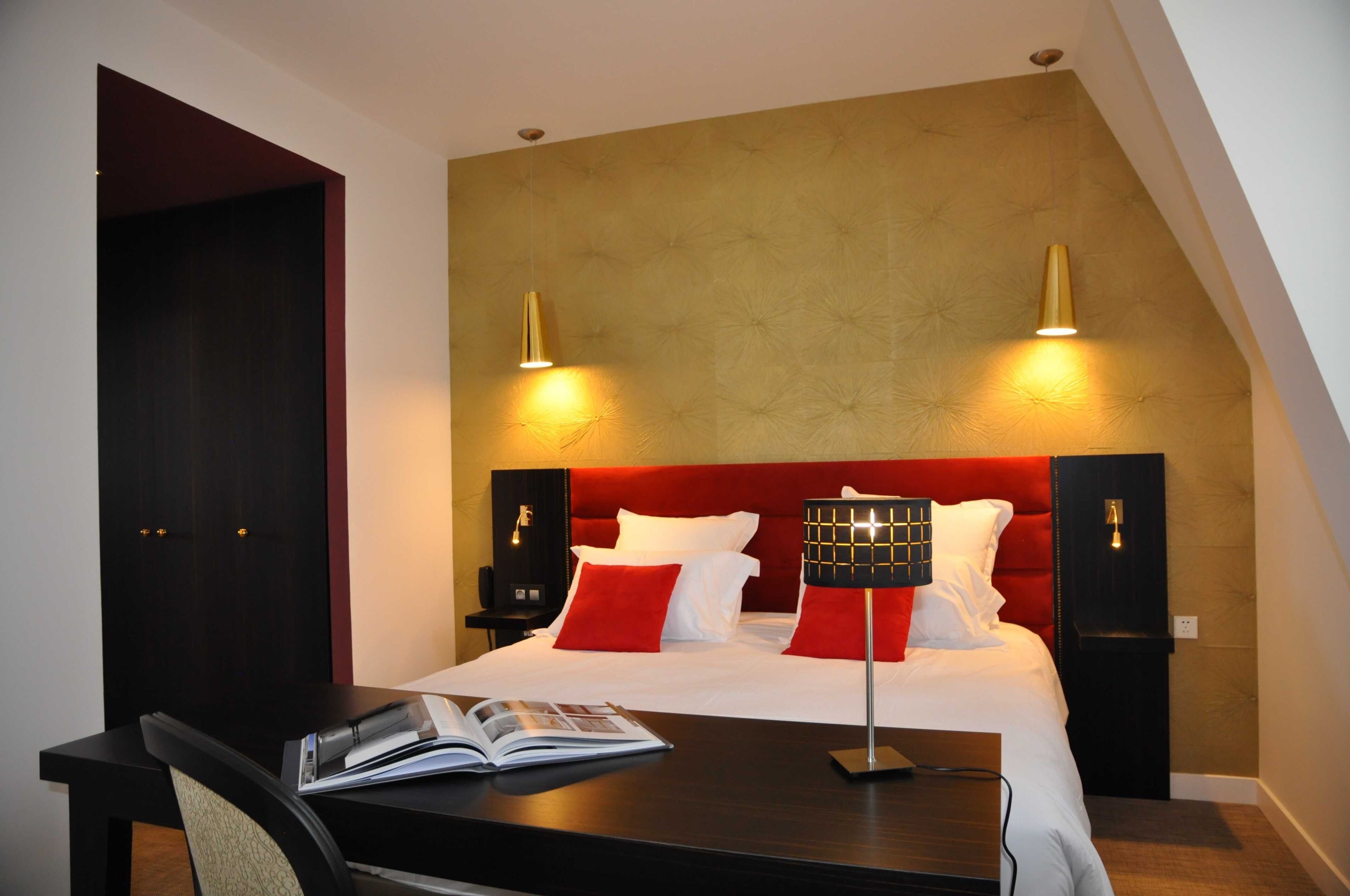 A hotel room with a red headboard, two bedside lamps, a desk with a chair, and a book on the desk.