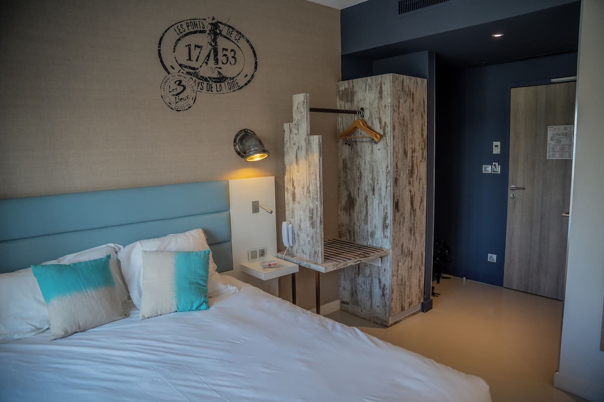 Una habitación de hotel con una cama, un armario de madera, una mesita de noche con una lámpara y un reloj de pared.