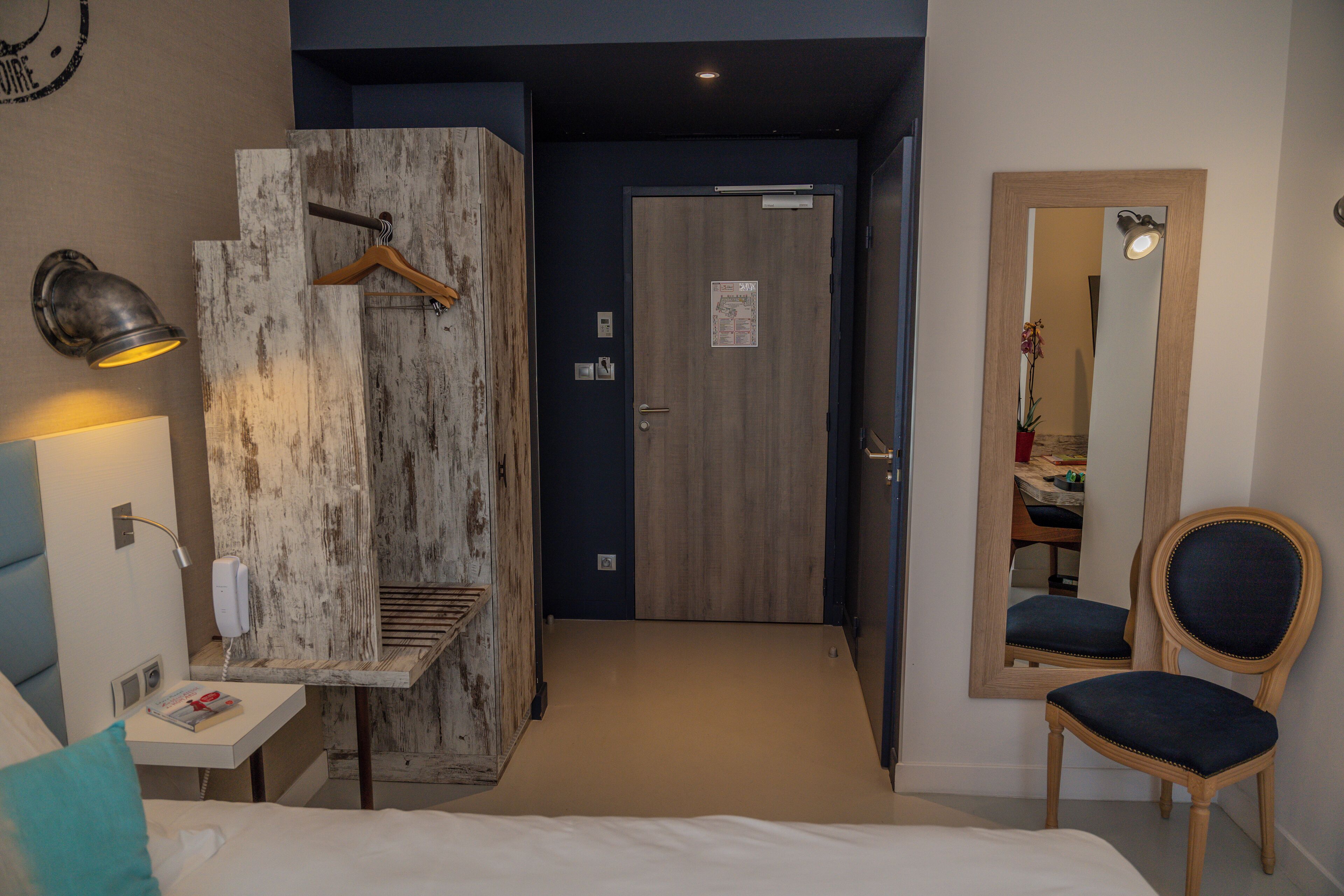 Habitación de hotel con cama, mesita de noche, lámpara, panel de pared de madera, puerta, espejo y una silla.