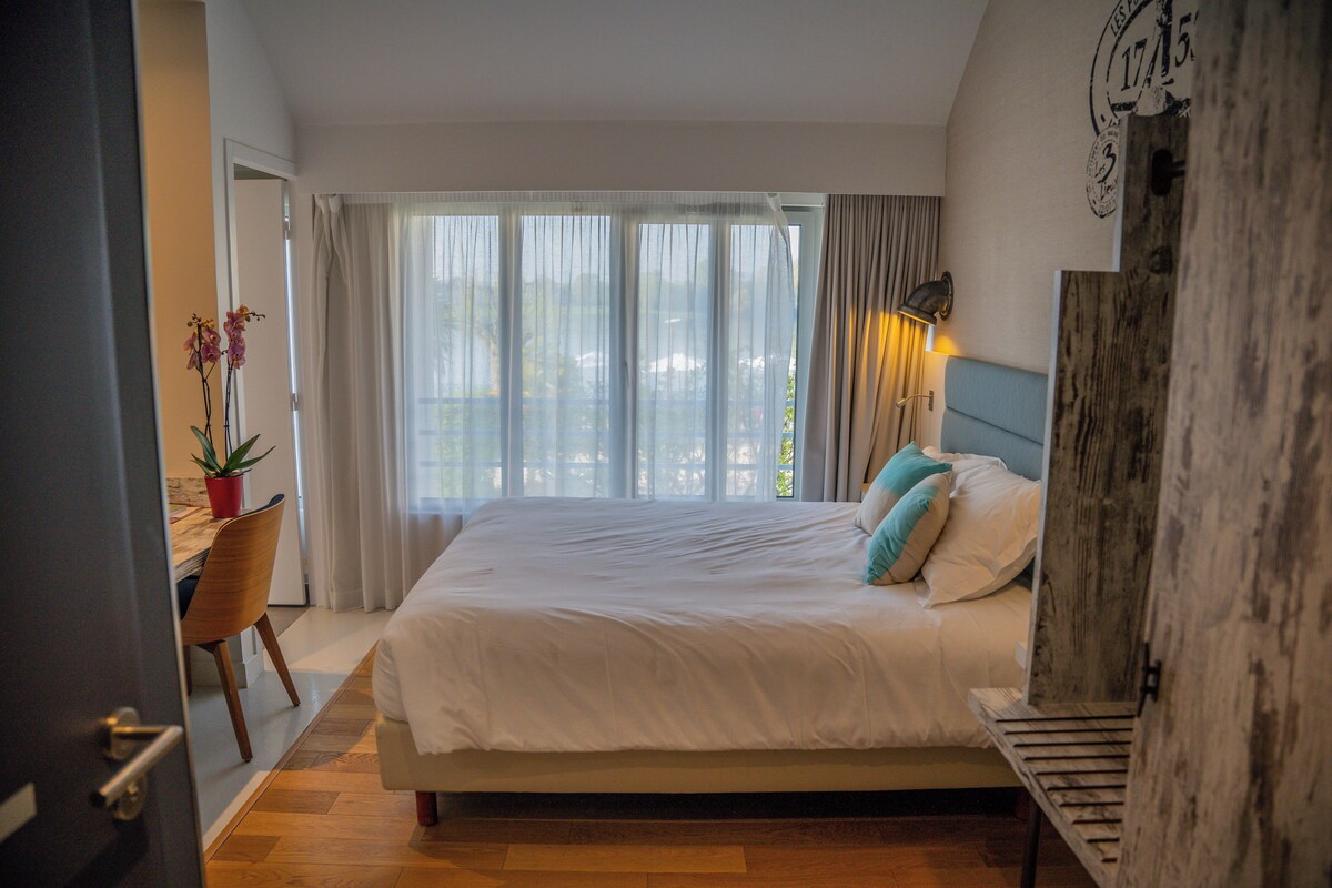 Un dormitorio con una cama grande, un escritorio con una silla, una planta en maceta y vistas al exterior a través de cortinas translúcidas.