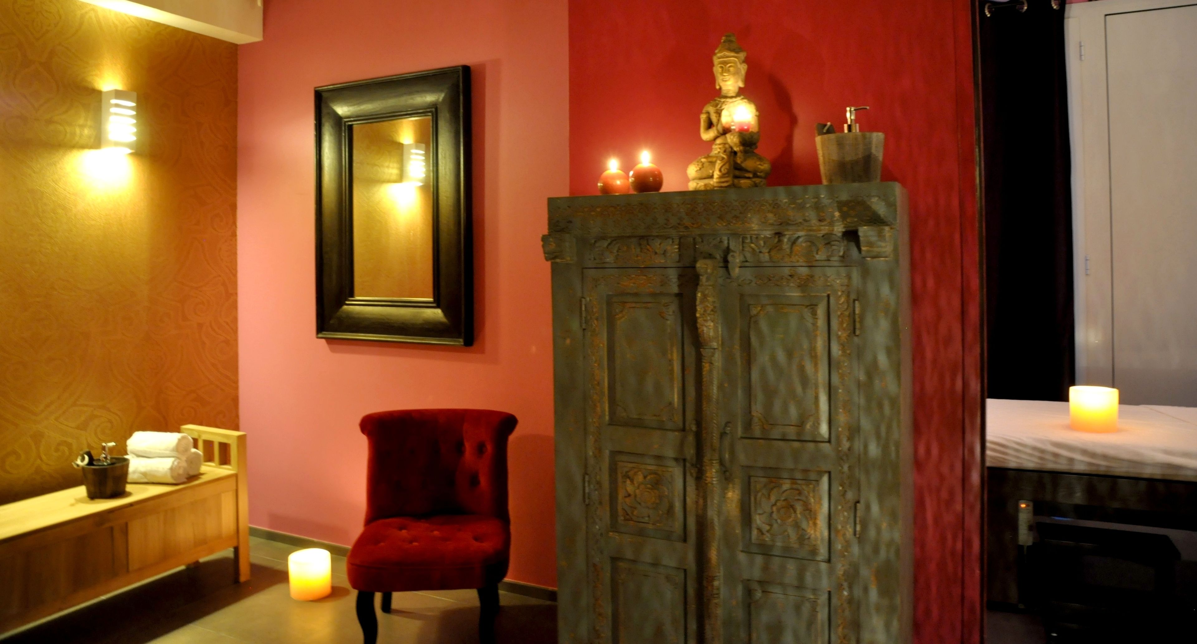 A room with a mirror, a red chair, a cabinet with a Buddha statue, and candles.