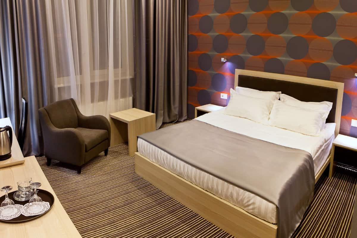 A hotel room with a bed, a chair, a desk with a kettle, and a tray with glasses.