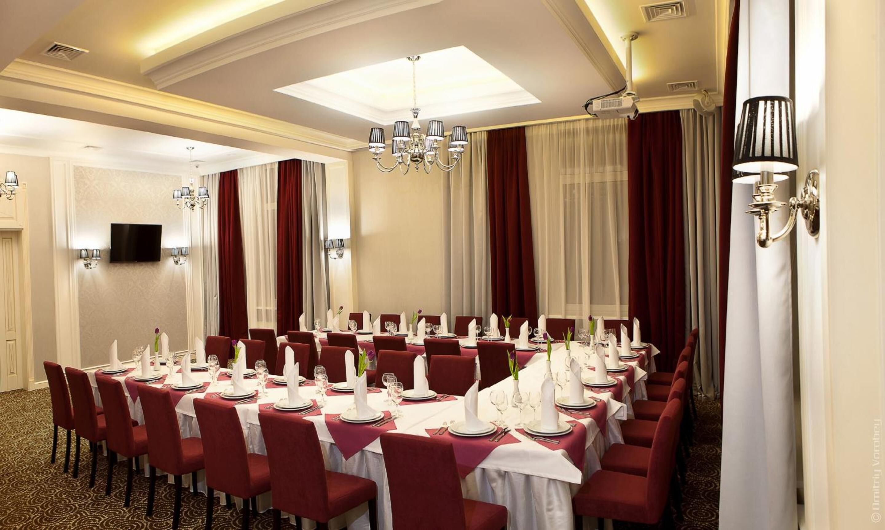 A banquet hall with round tables set for dining, a chandelier, and a television on the wall.