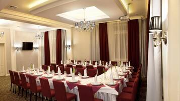 A banquet hall with round tables set for dining, a chandelier, and a television on the wall.