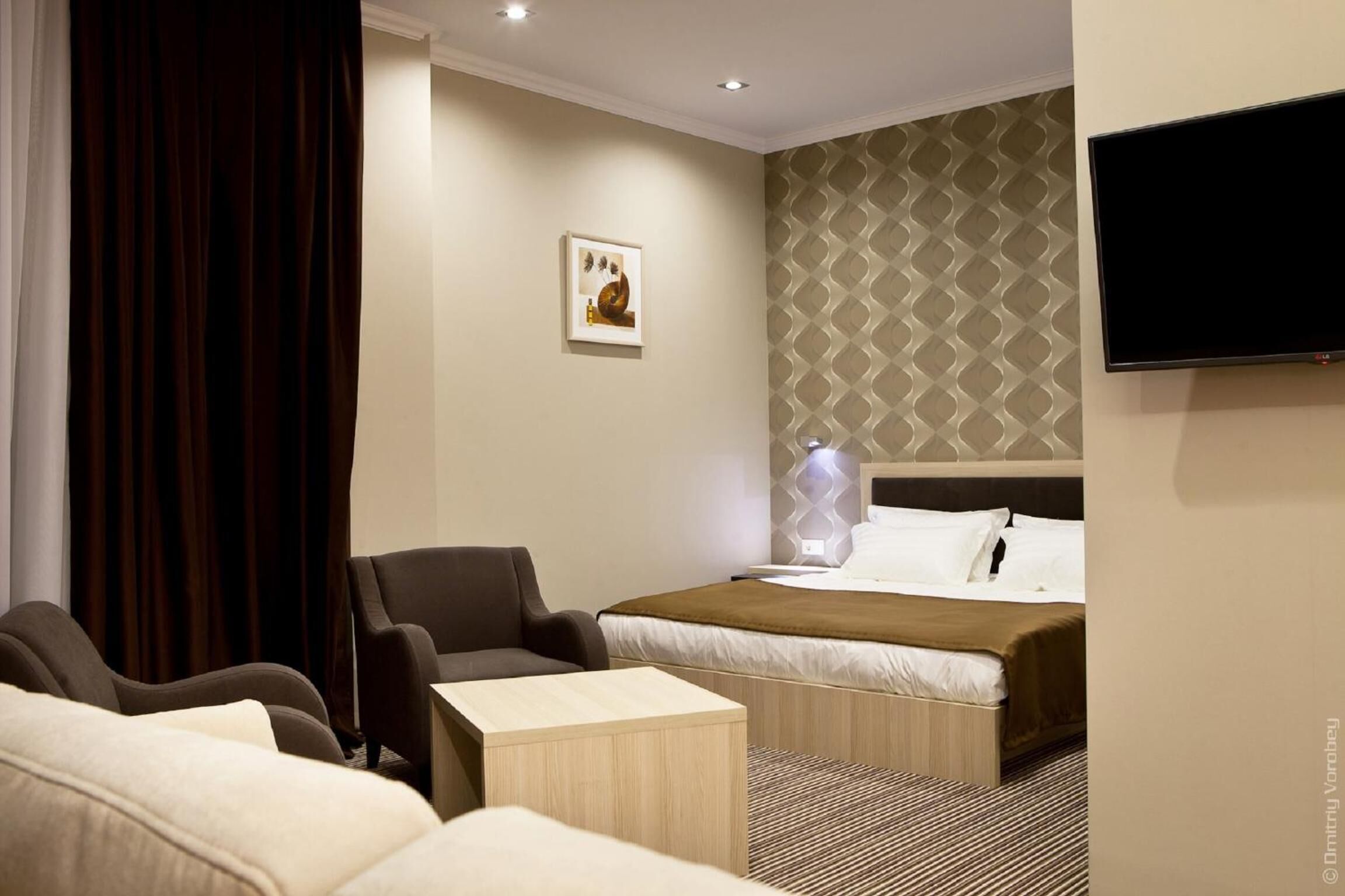 A hotel room with a bed, two armchairs, a small table, and a TV on the wall.