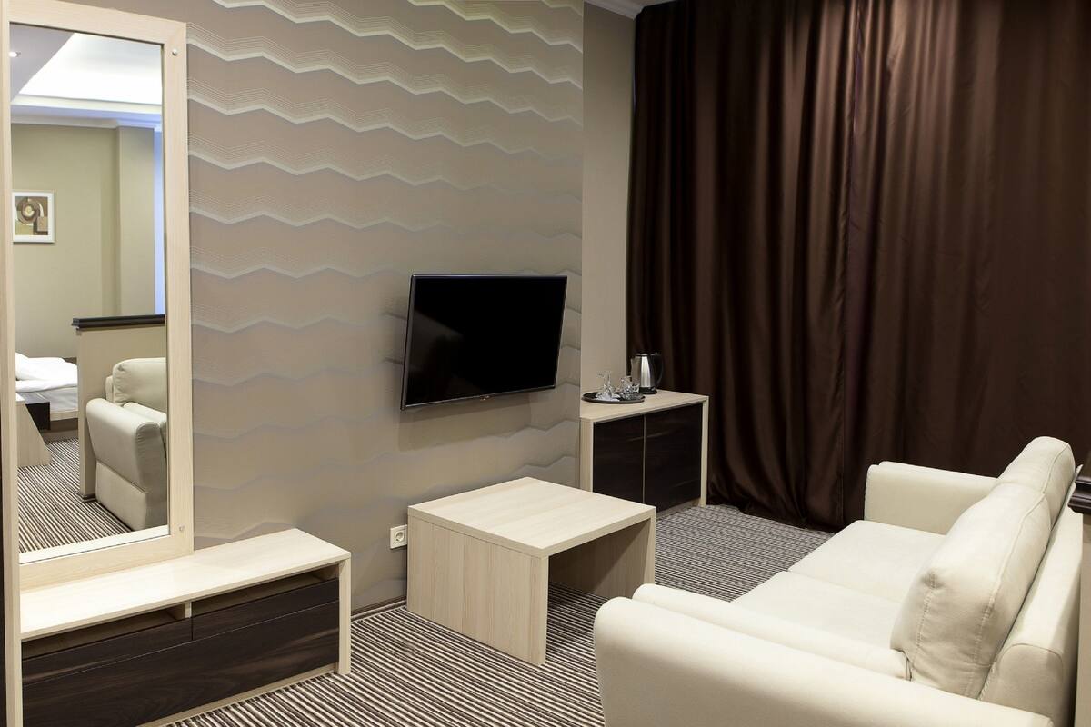 A modern hotel room with a large mirror, a flat-screen TV mounted on the wall, a small wooden side table, a white sofa, and a patterned carpet.