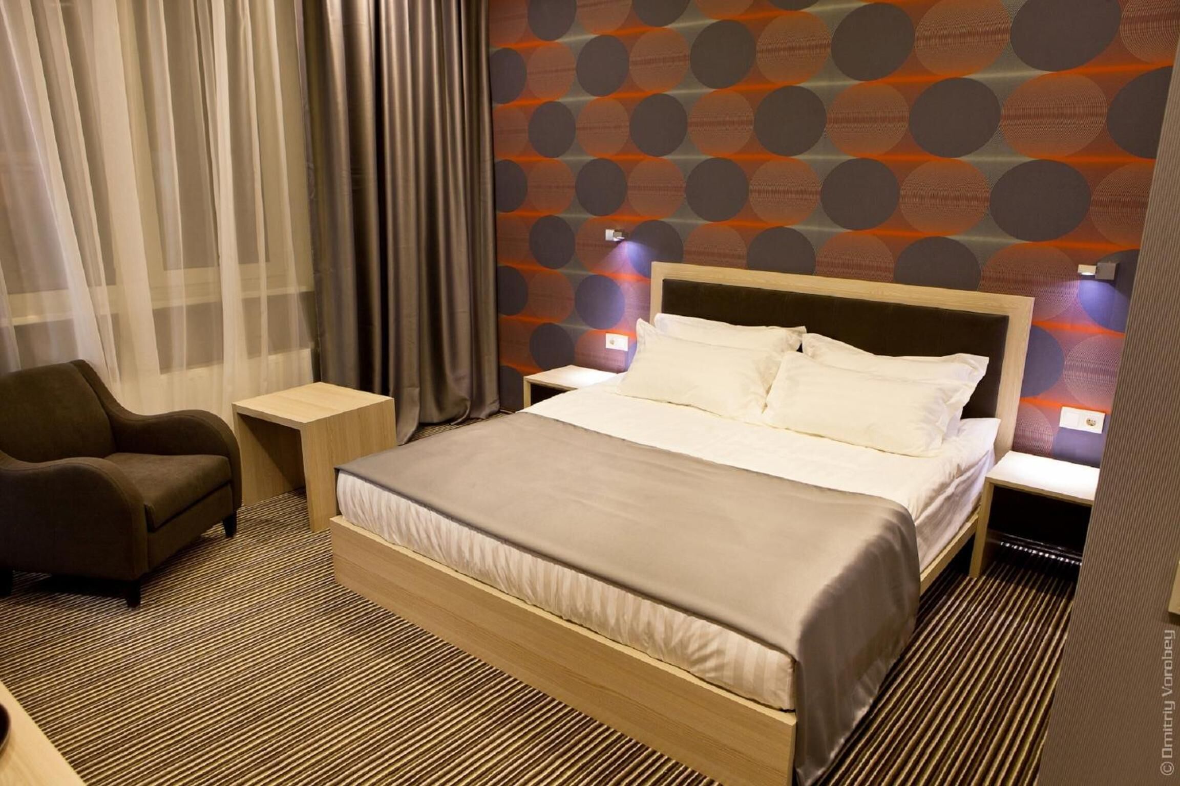 A hotel room with a bed, a nightstand, a chair, and a wall with a circular pattern.