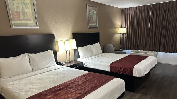 A hotel room with two beds, a nightstand with a lamp, a chair, and a window with curtains. - Rodeway Inn Bushnell (Bushnell)