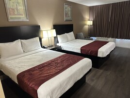 Habitación de hotel con dos camas, una mesita de noche con lámpara, una silla y un ventanal con cortinas.