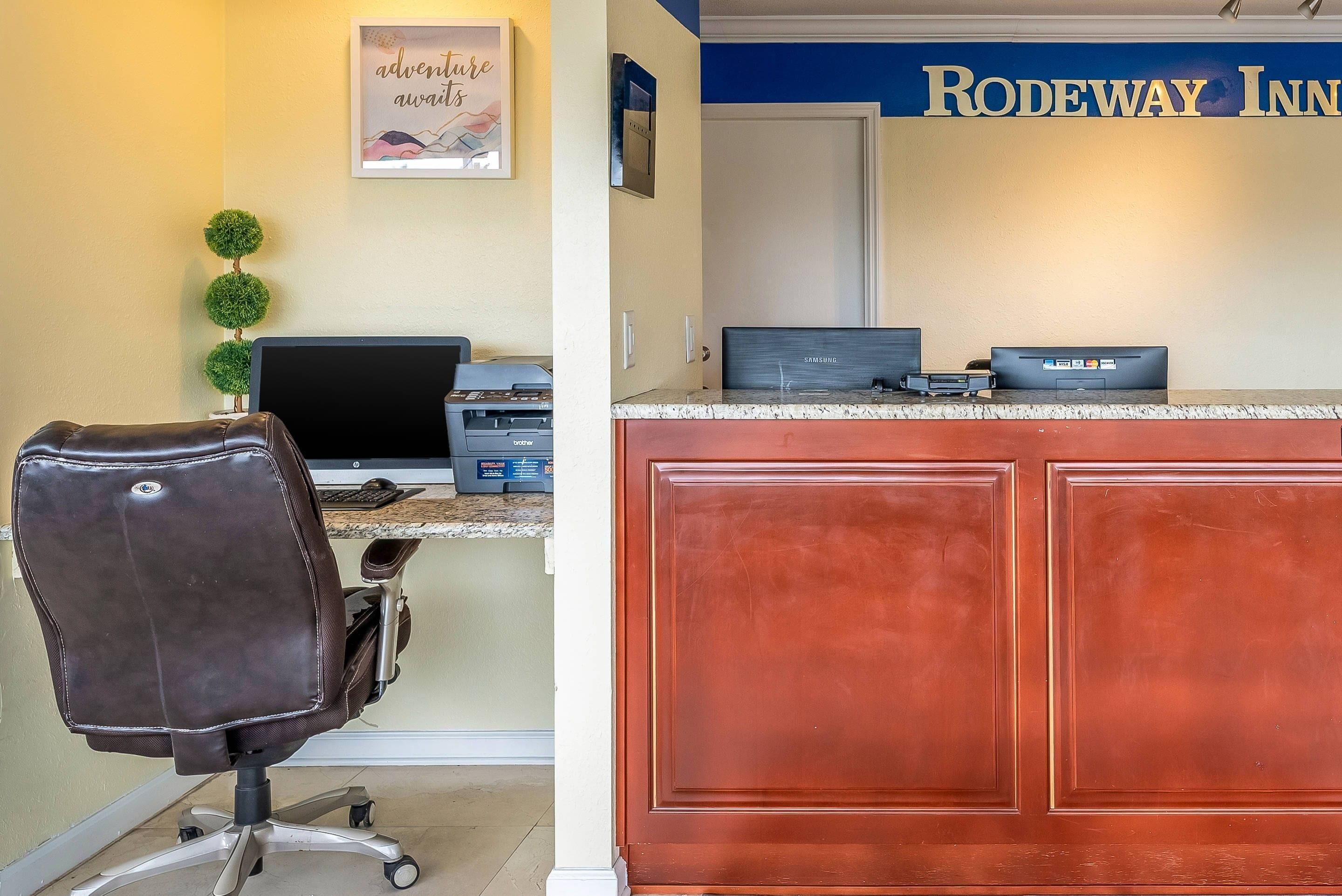 A hotel reception desk with a Samsung monitor, a printer, and a computer. There is a brown leather chair in front of the desk. The wall behind the desk has a sign that reads "RODEWAY INN."