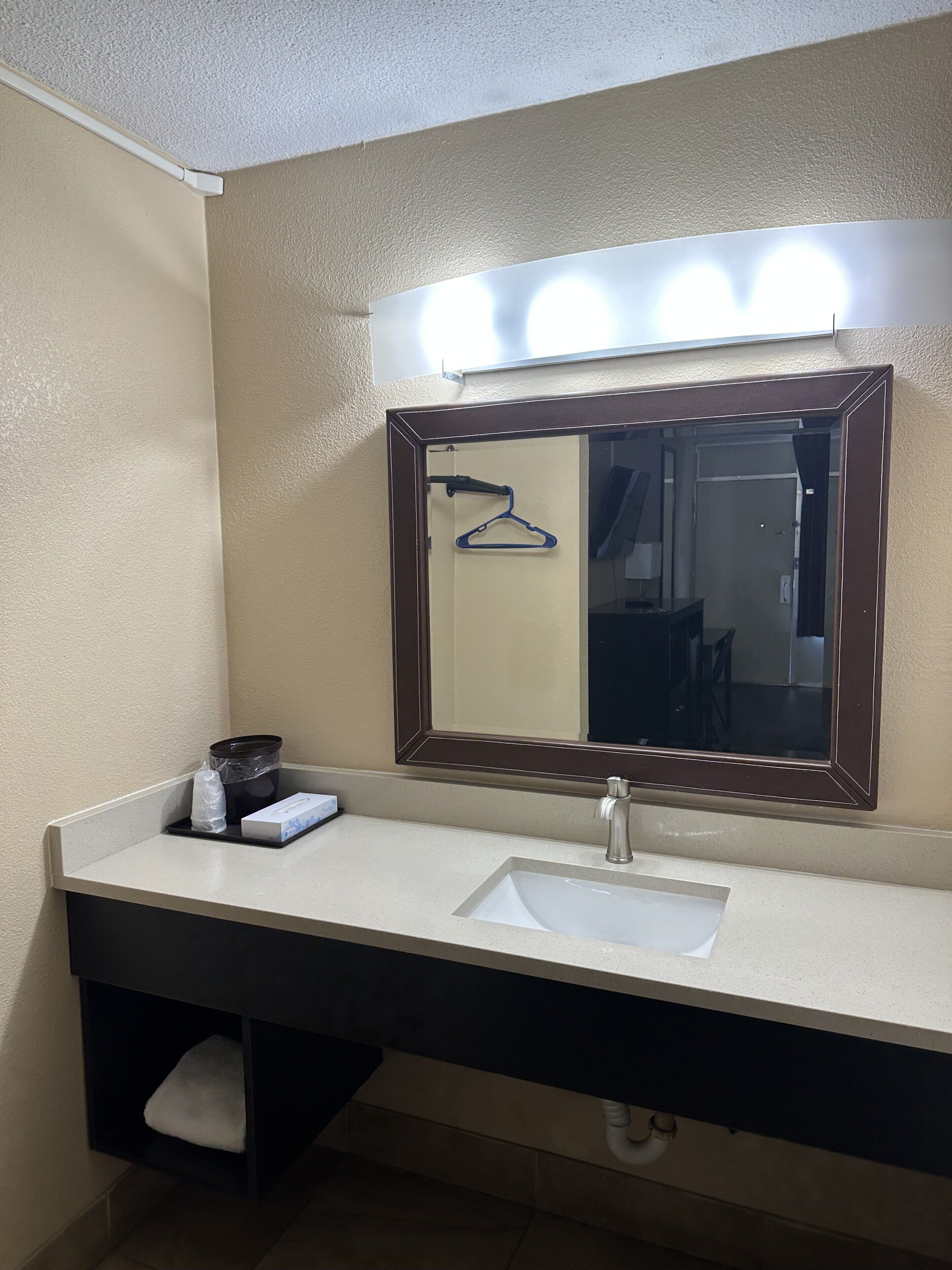 A bathroom with a white sink, a mirror with lights, a towel rack, and a trash bin.