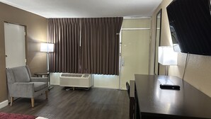 A hotel room with a bed, a desk, a chair, a TV, a lamp, a window with curtains, and an air conditioning unit. - Rodeway Inn Bushnell (Bushnell)