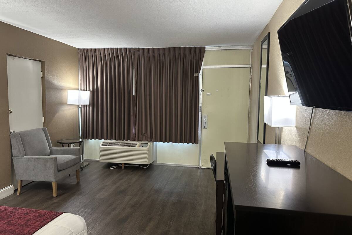 A hotel room with a bed, a desk, a chair, a TV, a lamp, a window with curtains, and an air conditioning unit.