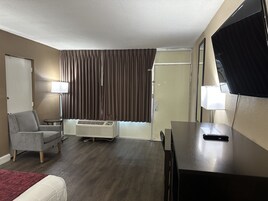Una habitación de hotel con cama, escritorio, silla, televisor, lámpara, ventana con cortinas y un equipo de aire acondicionado.