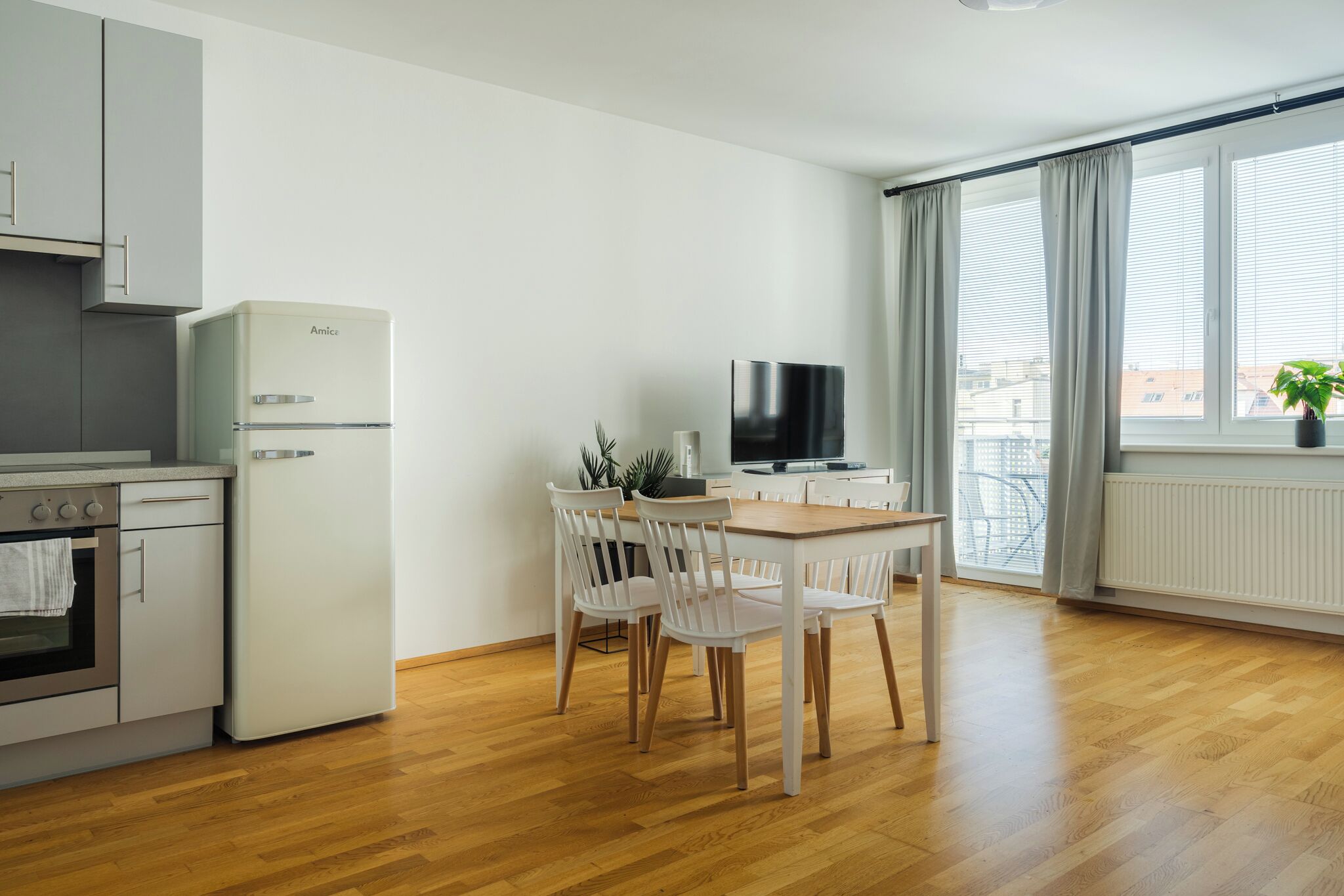 Una cocina moderna con electrodomésticos blancos, un comedor con mesa de madera y sillas blancas, y un amplio ventanal con cortinas.