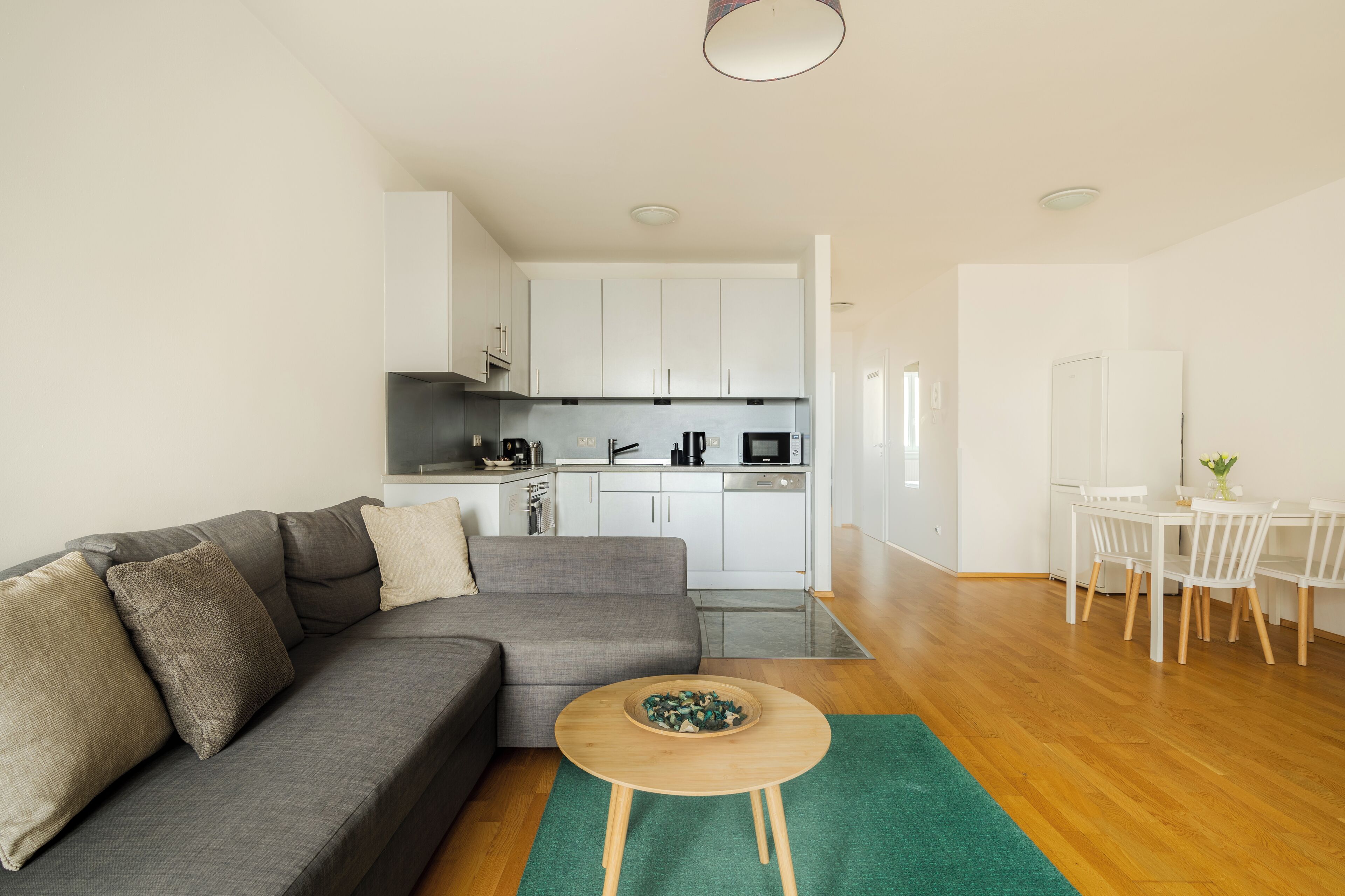 A modern living room with a grey sofa, a small wooden table, and a dining area with a white table and chairs.