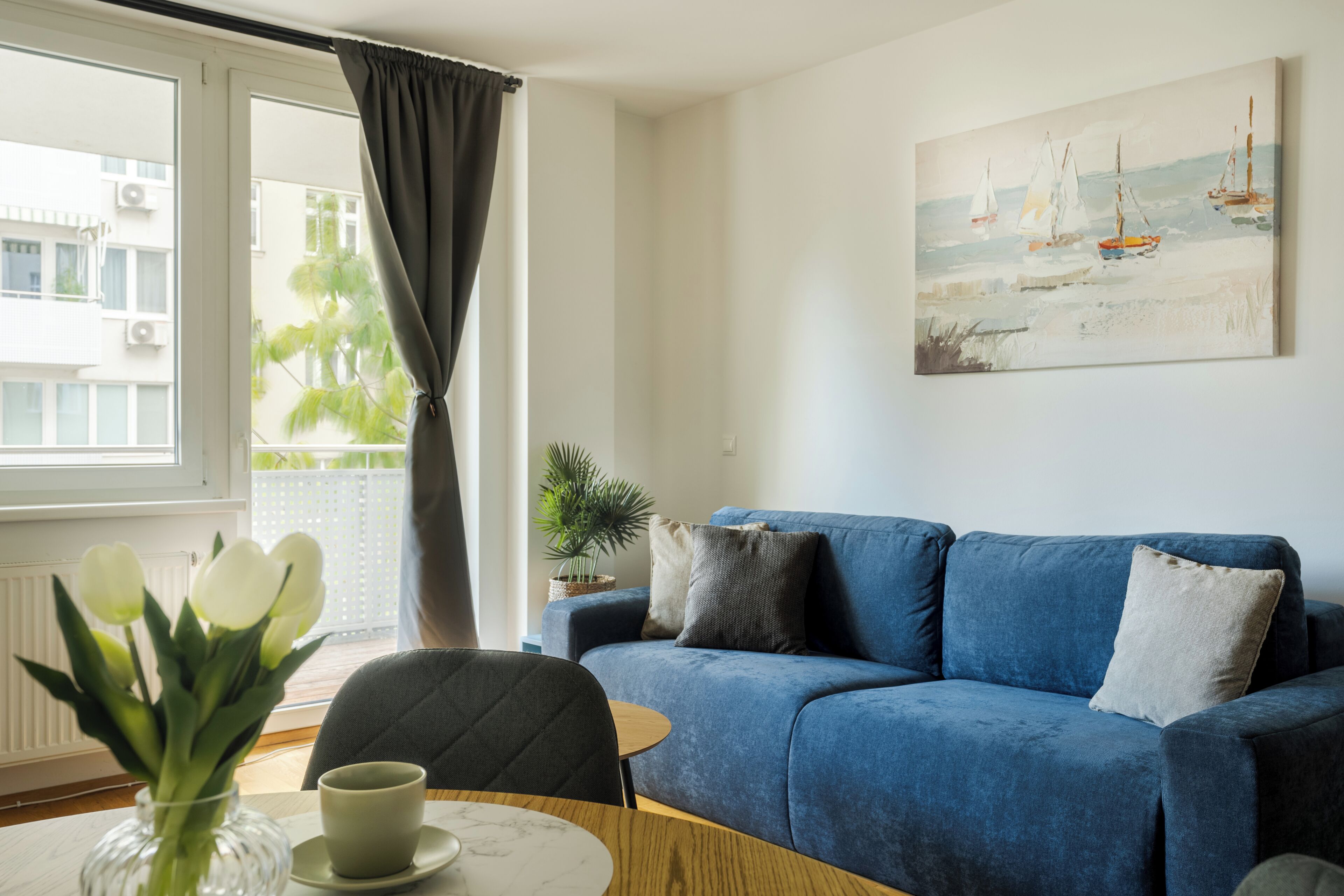 Un salon avec un canapé bleu, un tableau représentant des voiliers, une fenêtre avec des rideaux et un vase de fleurs posé sur une table.