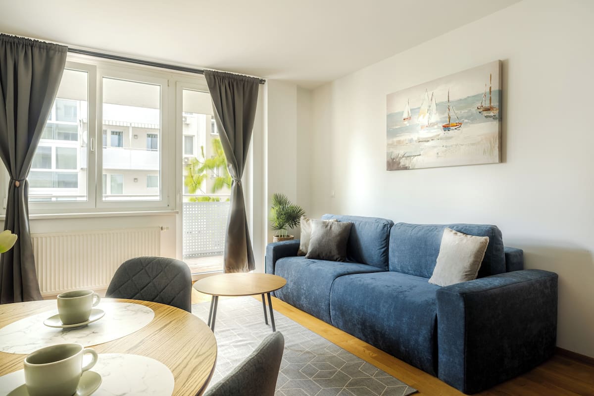 A modern living room with a blue sofa, a small round table with two cups, and a view of the outside through large windows.