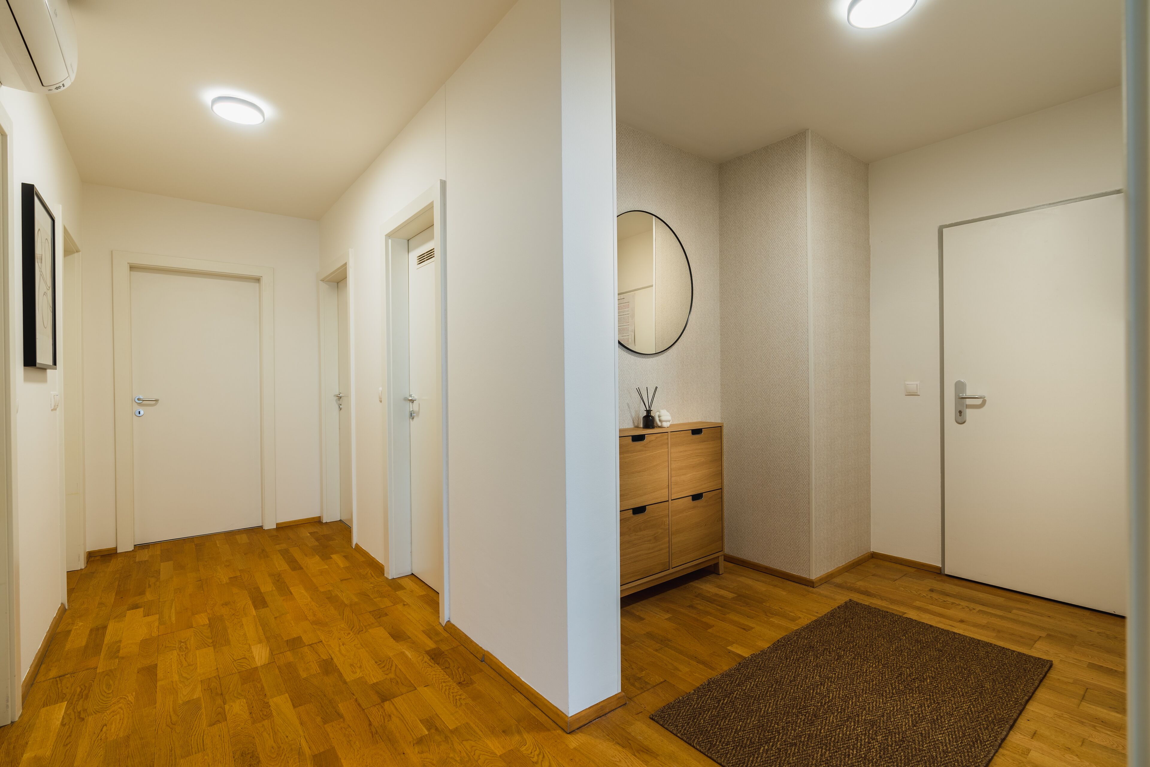 Un pasillo con piso de madera, paredes blancas y una alfombra marrón. Se observan tres puertas y un mueble de madera con cajones.