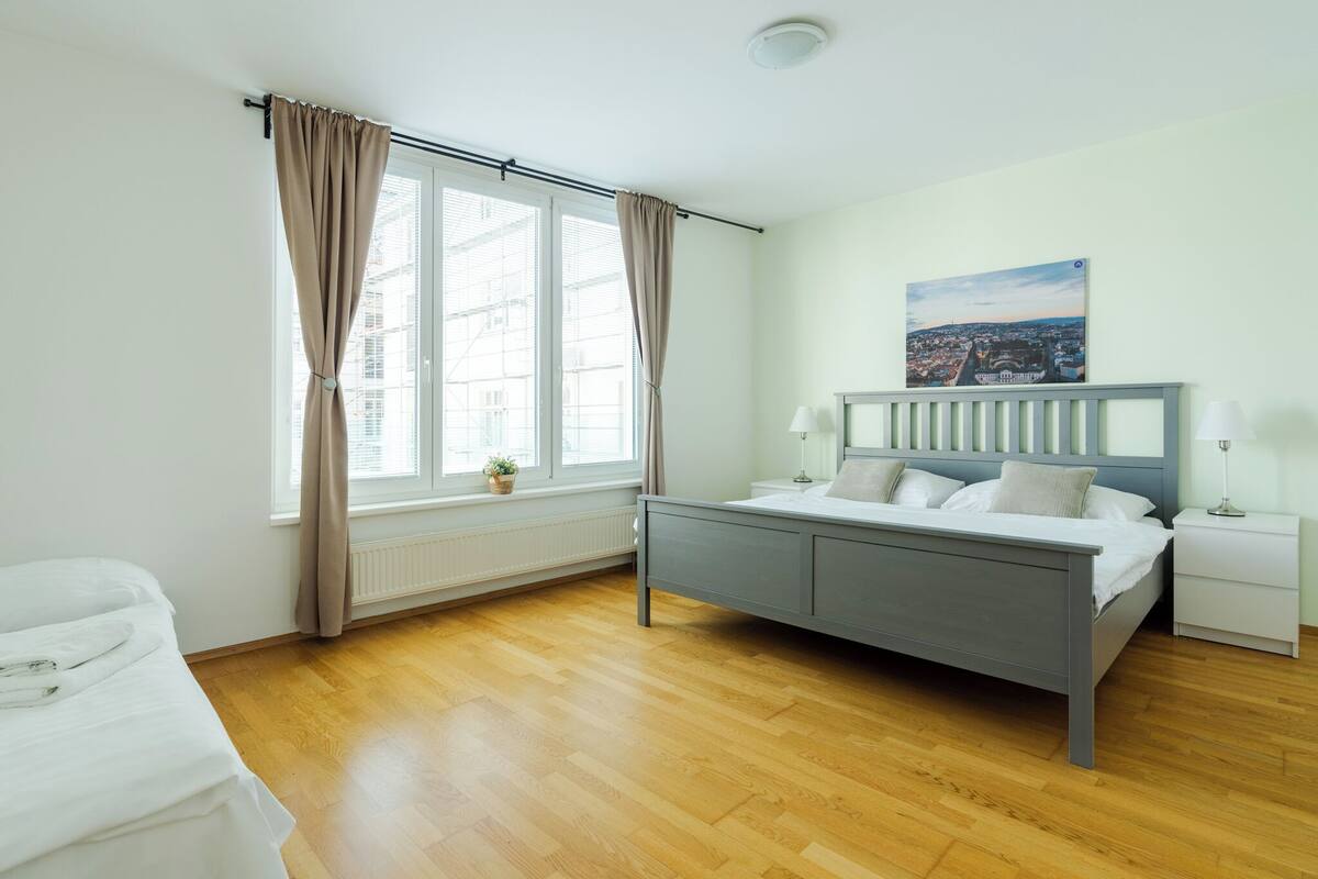 A bedroom with a bed, a nightstand, a lamp, a window with curtains, and a cityscape painting on the wall.