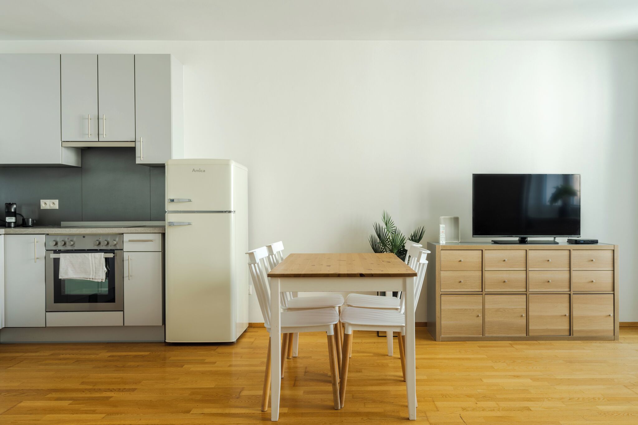 Une cuisine moderne avec des meubles blancs, une table à manger en bois et un téléviseur posé sur un meuble.