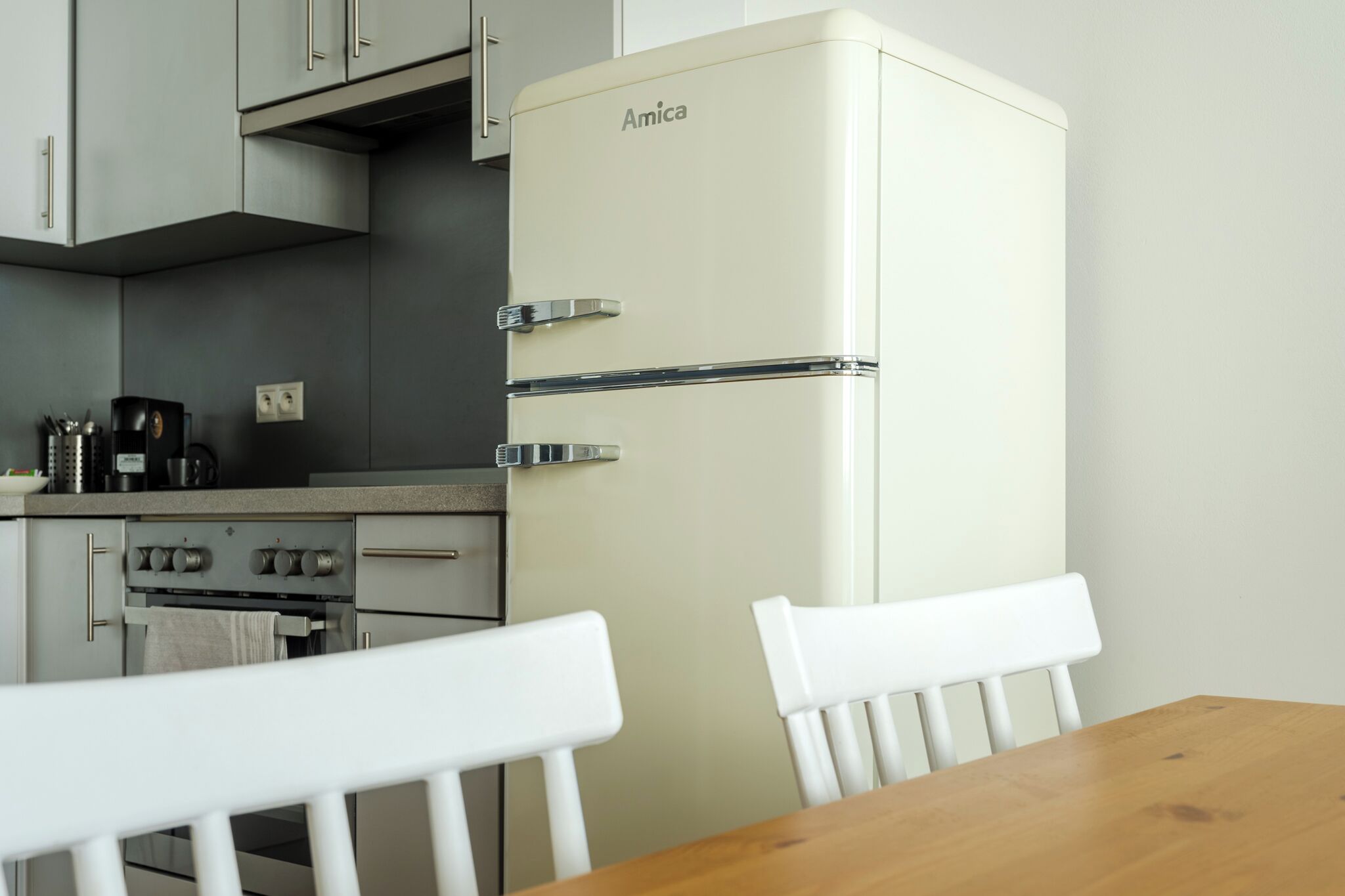 A kitchen with a white Amica refrigerator, a stove, and white chairs at a wooden table.