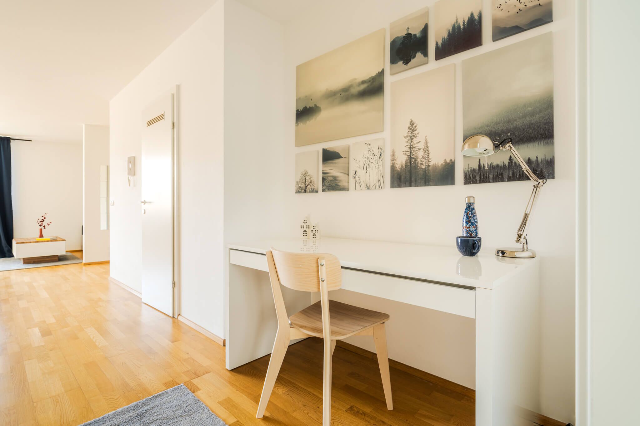 Une pièce moderne et minimaliste, dotée d’un bureau en verre, d’une chaise en bois et d’un mur orné de tableaux représentant des paysages.