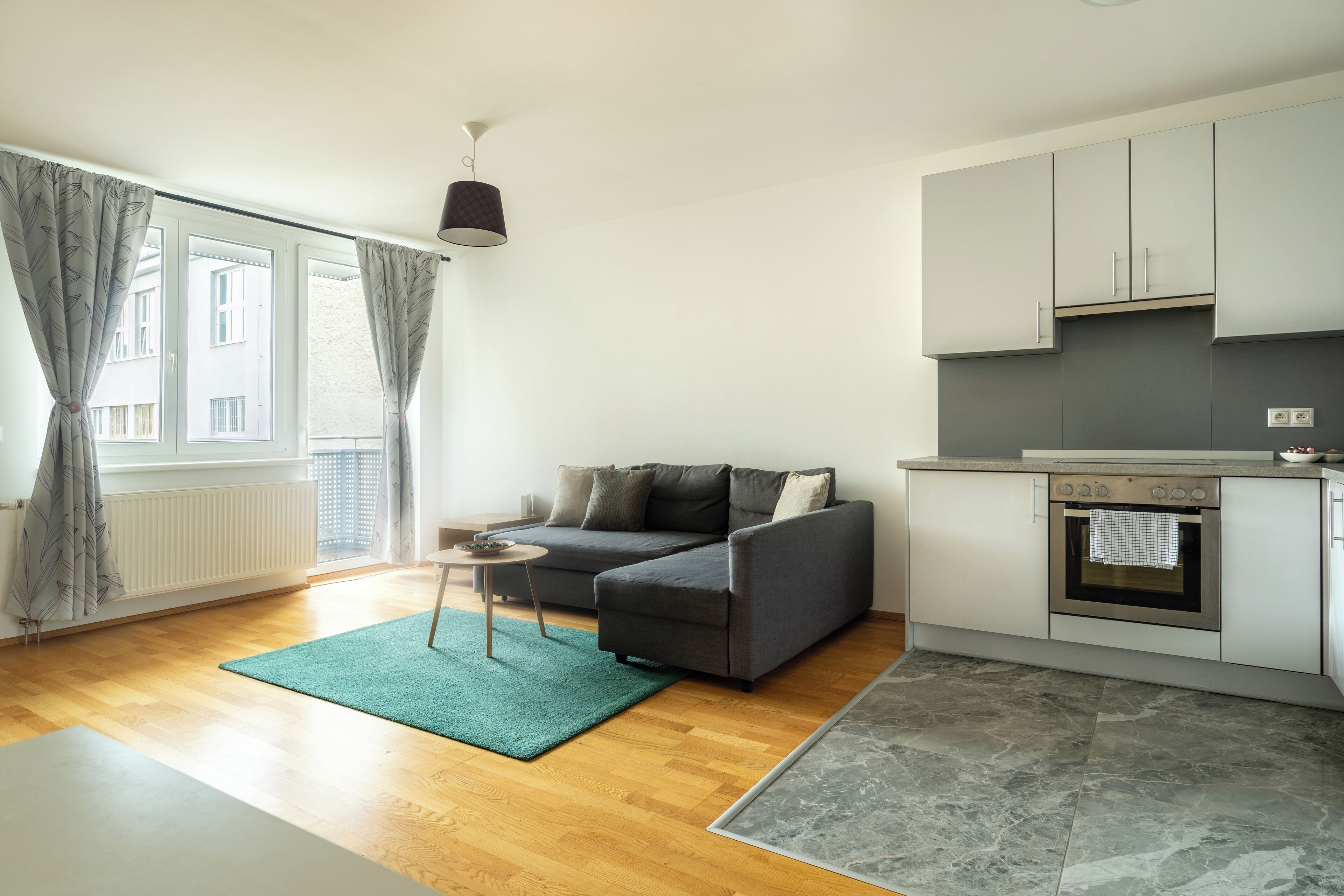 A modern living room with a grey sofa, a small side table, and a green rug. It features a kitchen with grey cabinets and a built-in oven.