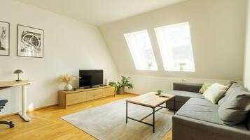 Una sala de estar moderna con un sofá, una mesa de centro, un televisor sobre un mueble de madera y un escritorio con una silla. Dos ventanas dejan entrar luz natural.