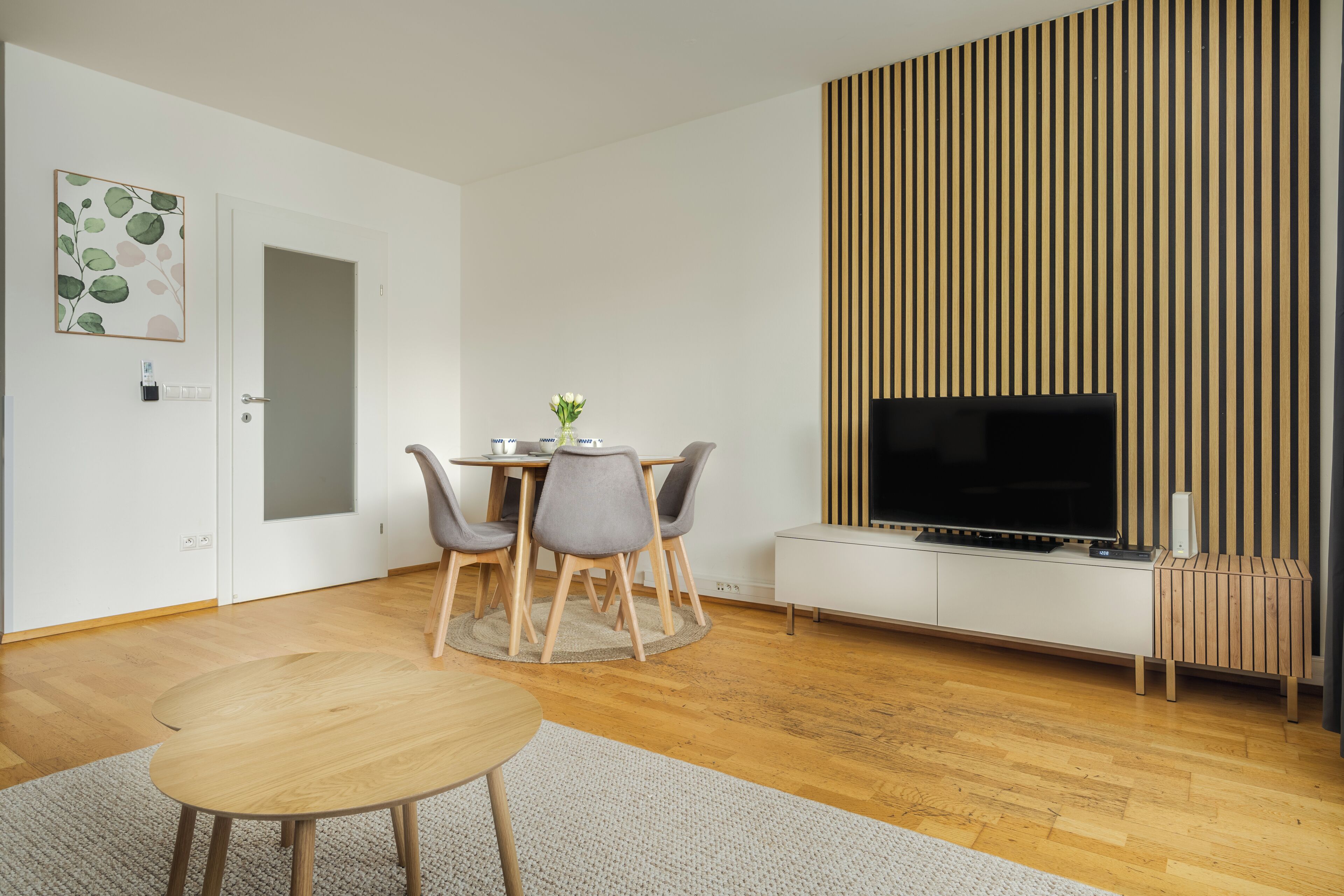 A modern living room with a wooden floor, a round dining table with chairs, a flat-screen TV mounted on a white cabinet, and a vertical wooden panel on the wall.