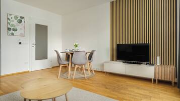 A modern living room with a wooden floor, a round dining table with chairs, a flat-screen TV mounted on a white cabinet, and a vertical wooden panel on the wall.