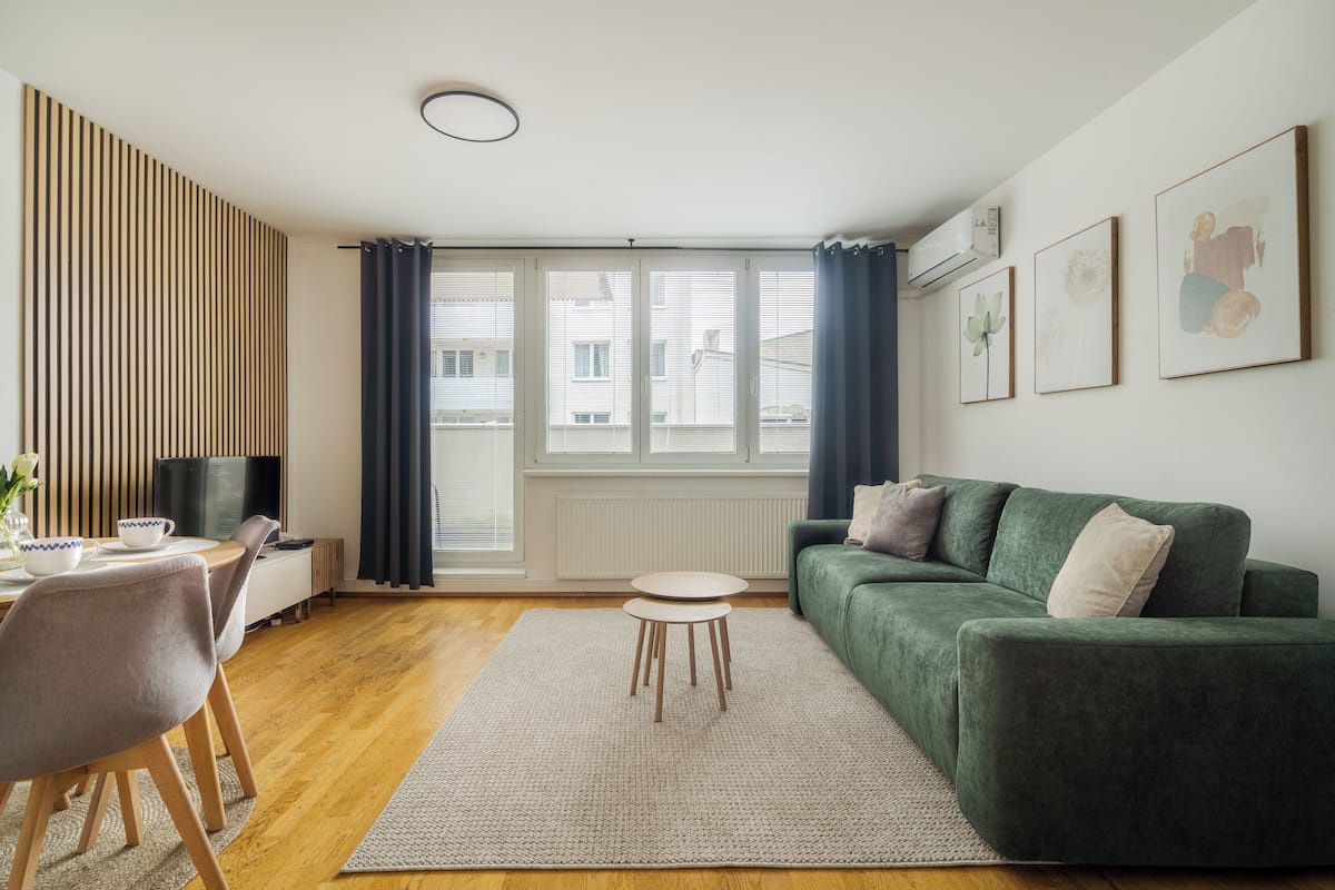 A modern living room with a green sofa, a dining table with chairs, a small round table, and a large window with curtains.
